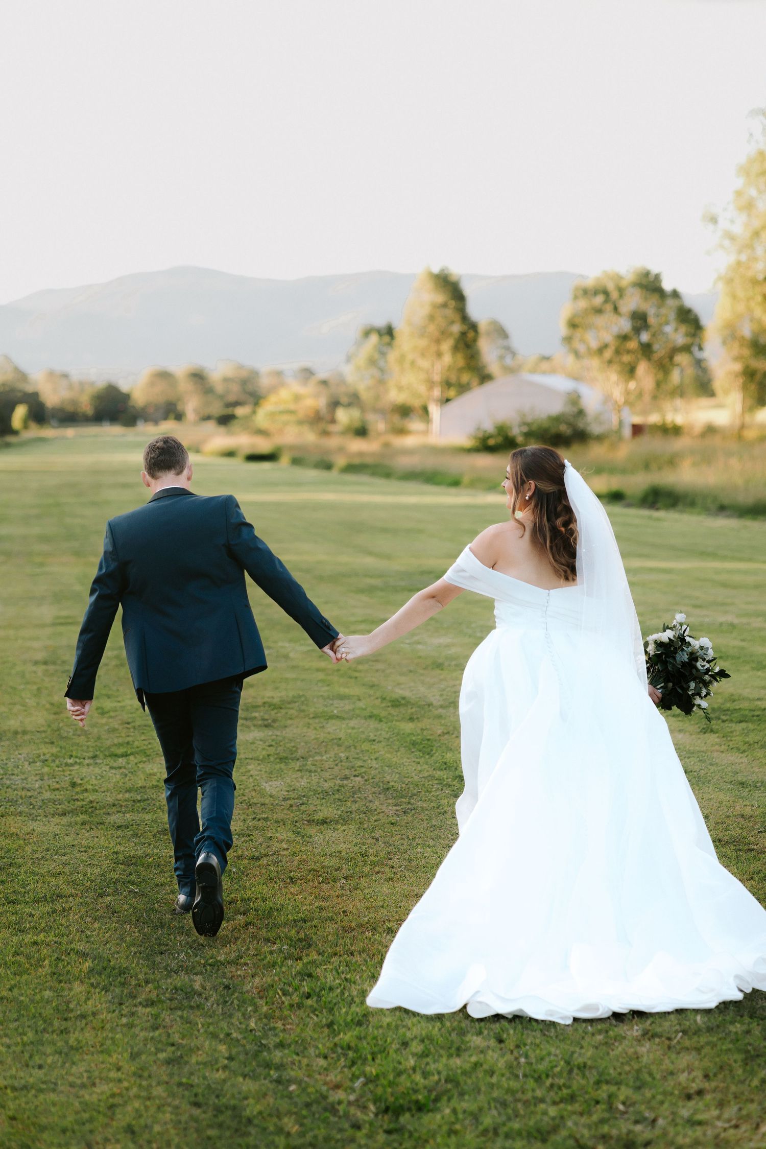 Bride and groom walking hand in hand across green field with mountain backdrop on their wedding day.