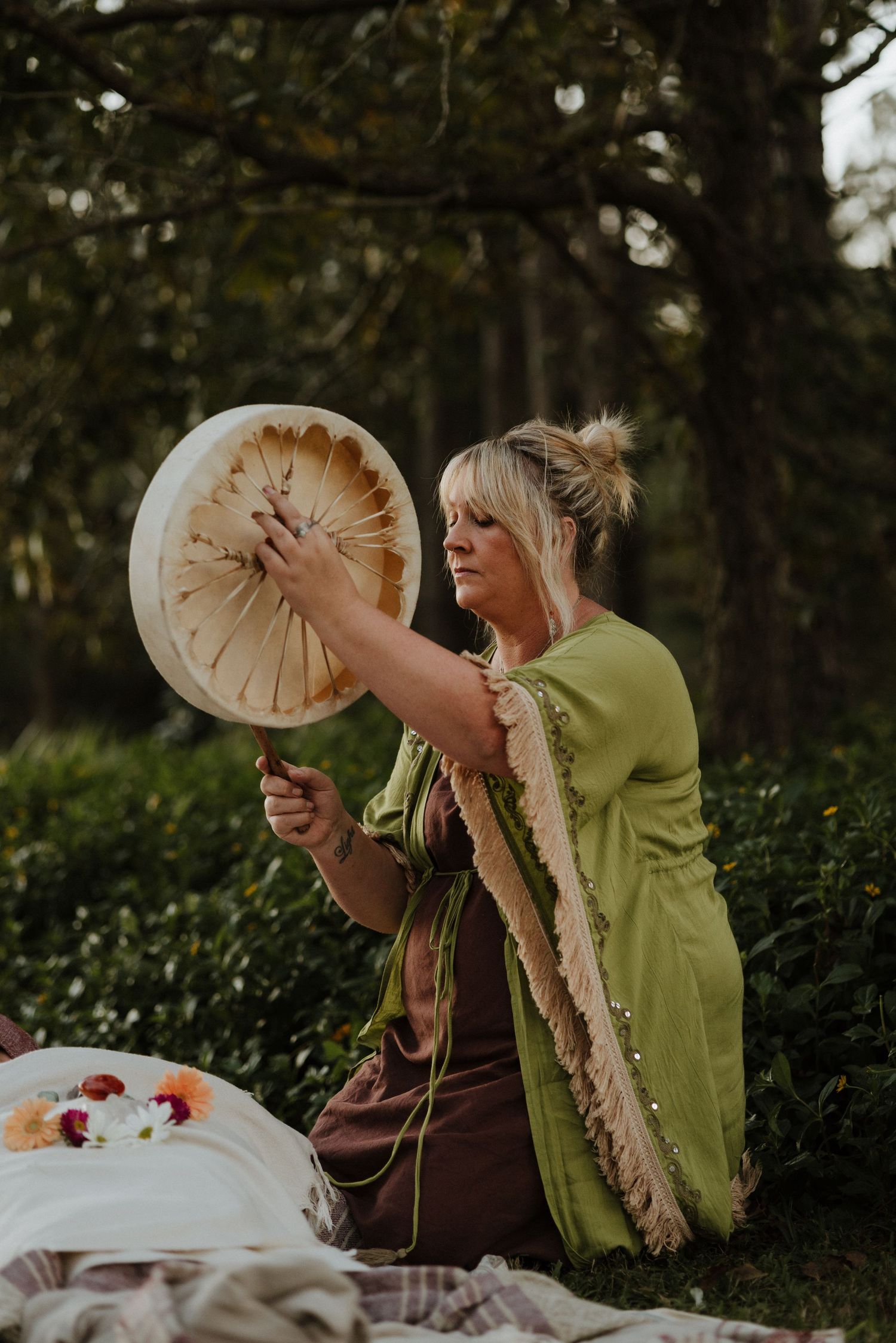 Woman in green shawl playing a frame drum outdoors in a lush garden setting.