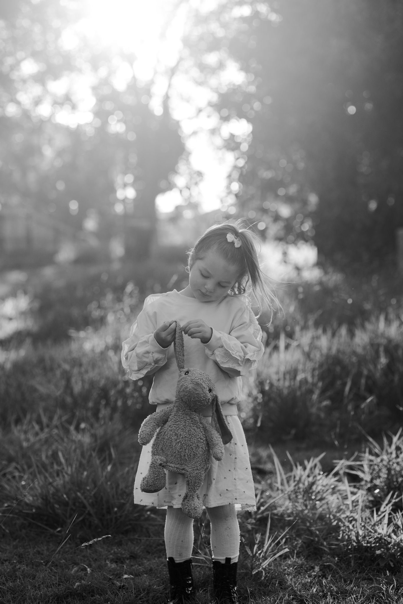 Black and white photo of a young girl in a dress holding a stuffed bunny toy outdoors in soft sunlight.