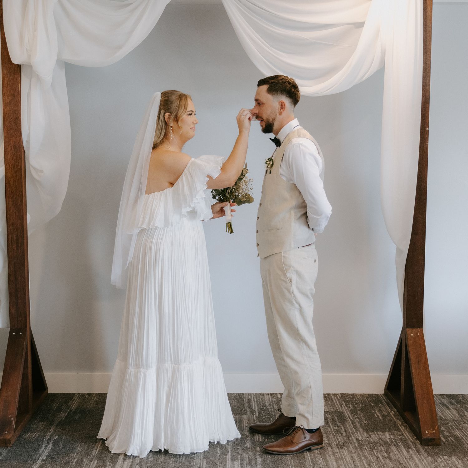 Bride and groom share a tender moment under a draped arch, exchanging vows in a boho-chic wedding ceremony.