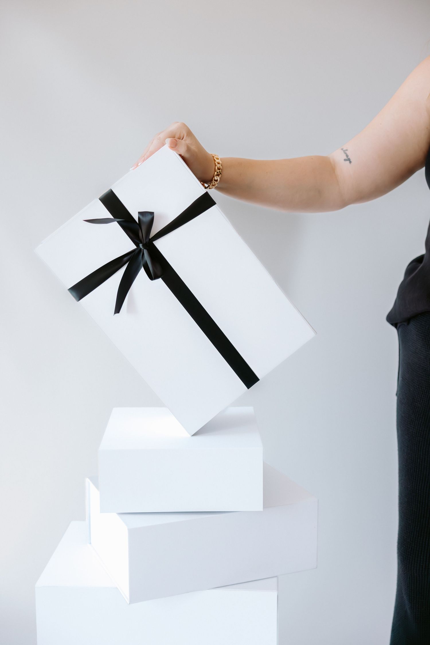 Person placing a white gift box with black ribbon bow on top of stacked white boxes against a light background.