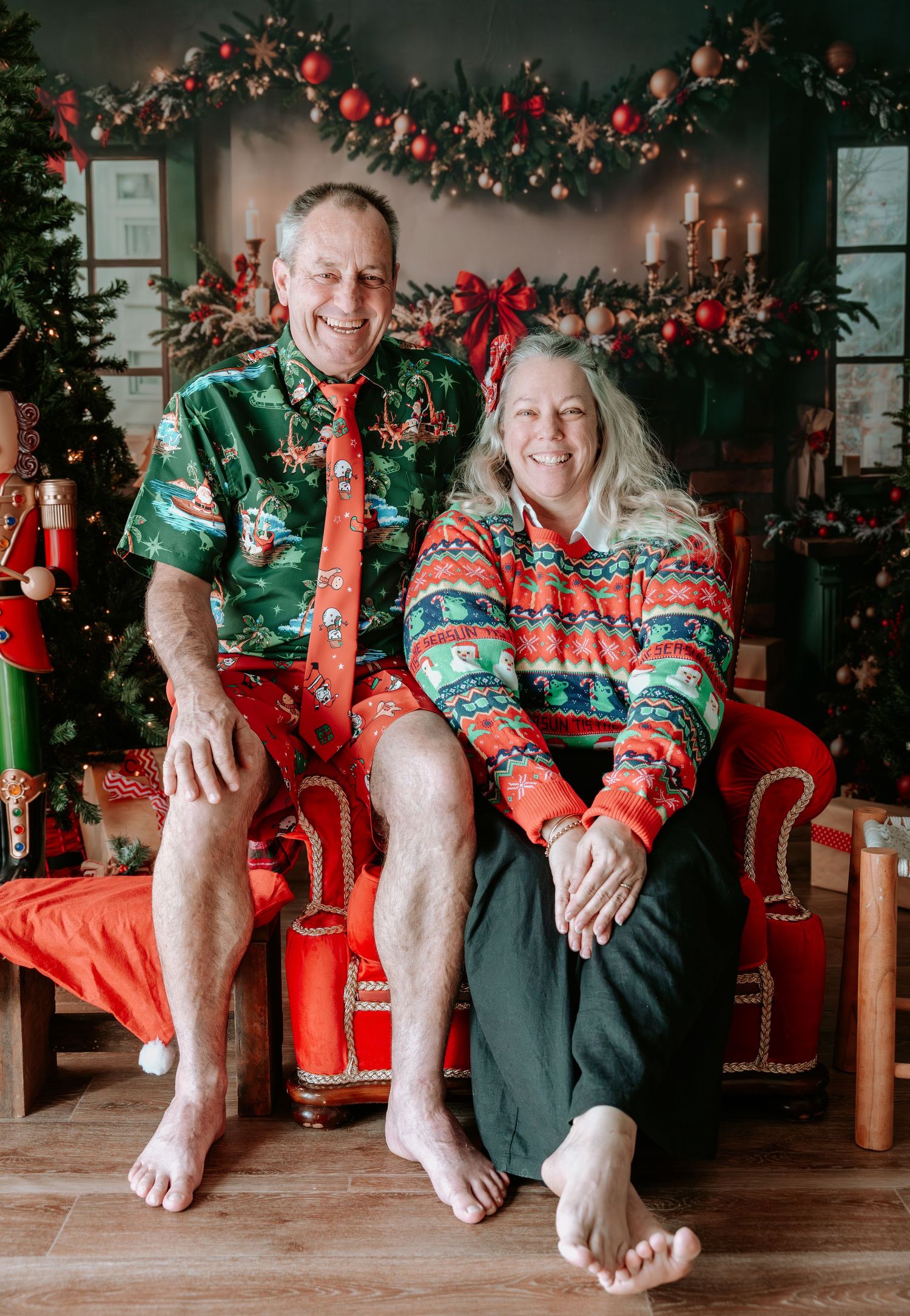 Smiling couple in festive Christmas sweater and Hawaiian shirt sitting barefoot on red chair with holiday decorations.