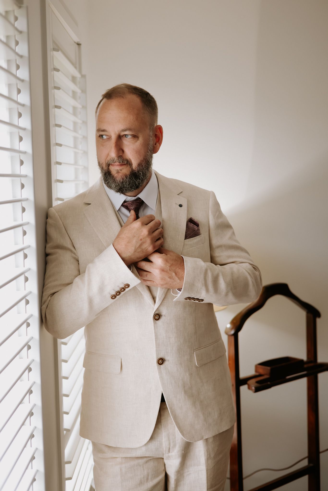 Middle-aged bearded man in beige linen suit adjusting tie near window with shutters.