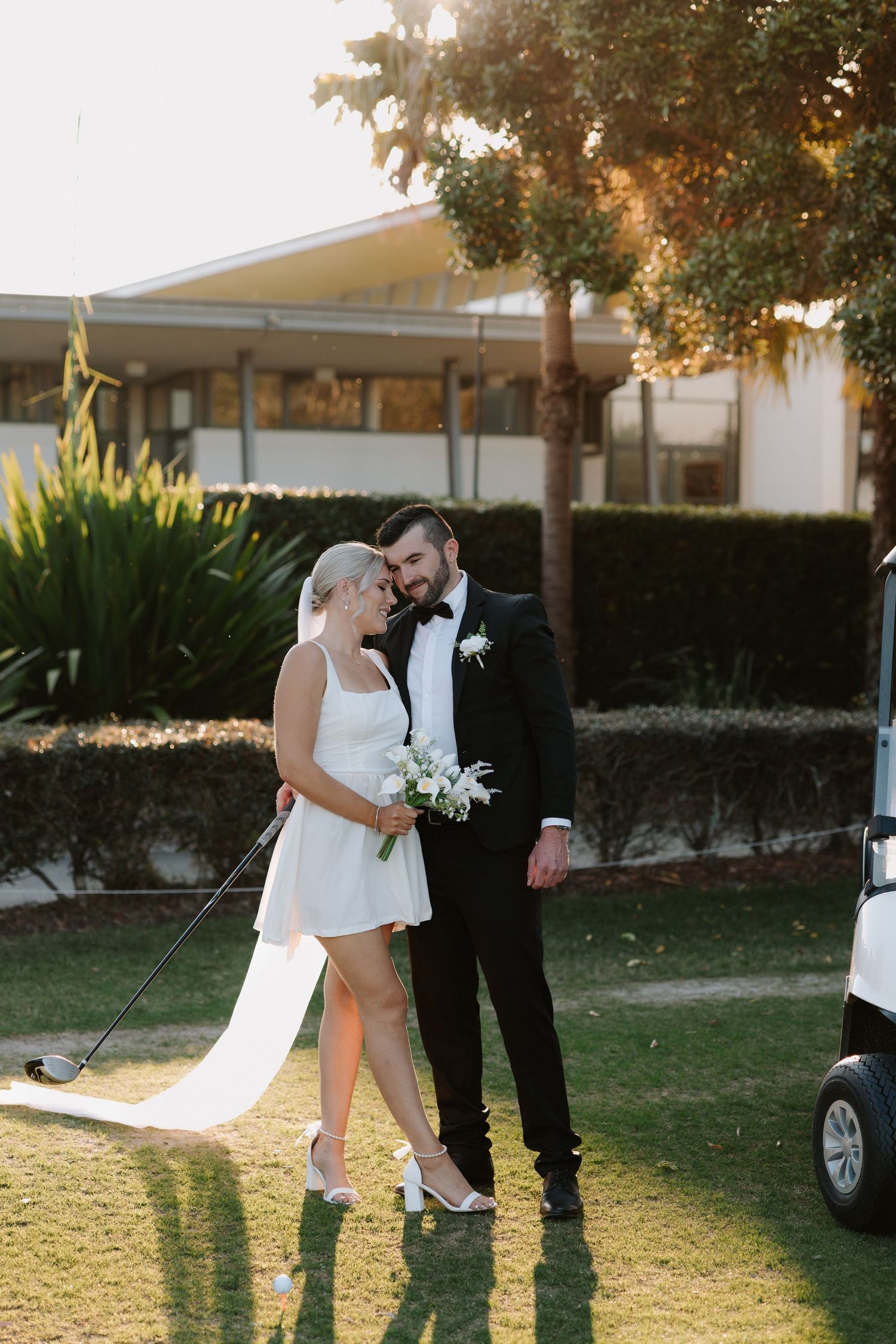 Bride in short white dress holding golf club and groom in black tuxedo pose together on golf course at sunset.