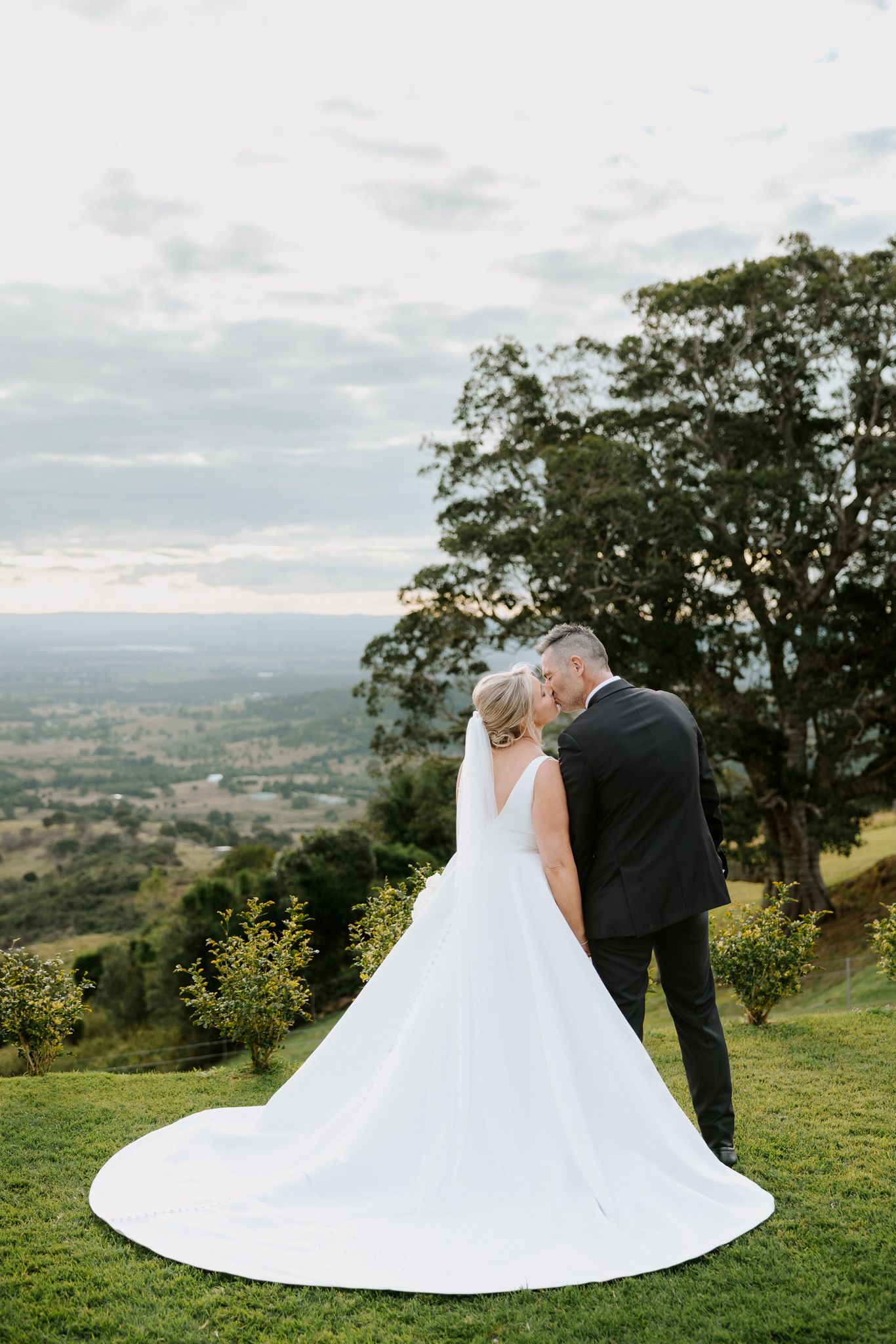 Bride and groom share a kiss overlooking a scenic valley landscape, bride wearing elegant white gown with long train.