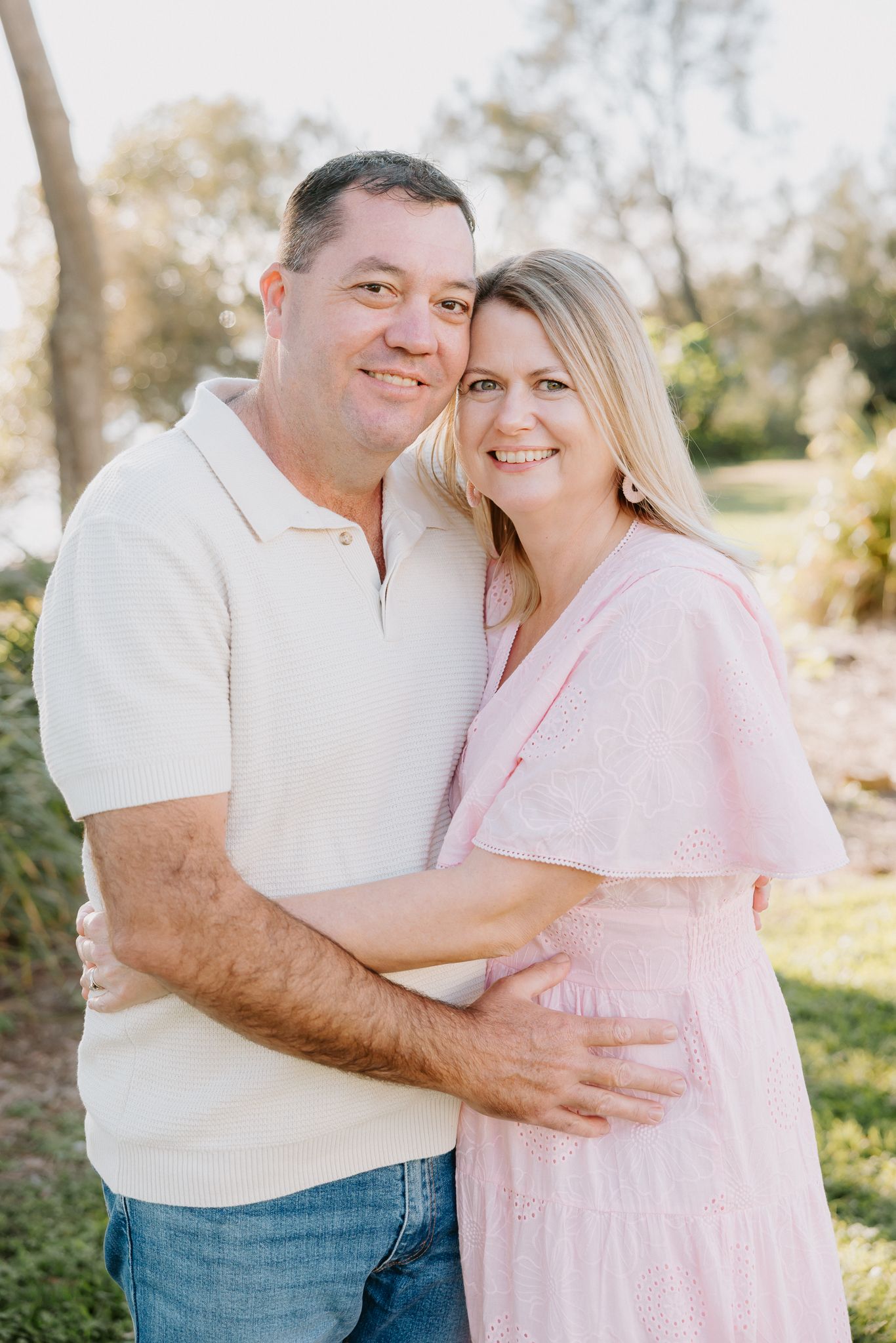A smiling couple embraces outdoors, woman in pink dress and man in cream polo shirt, with greenery in the background.