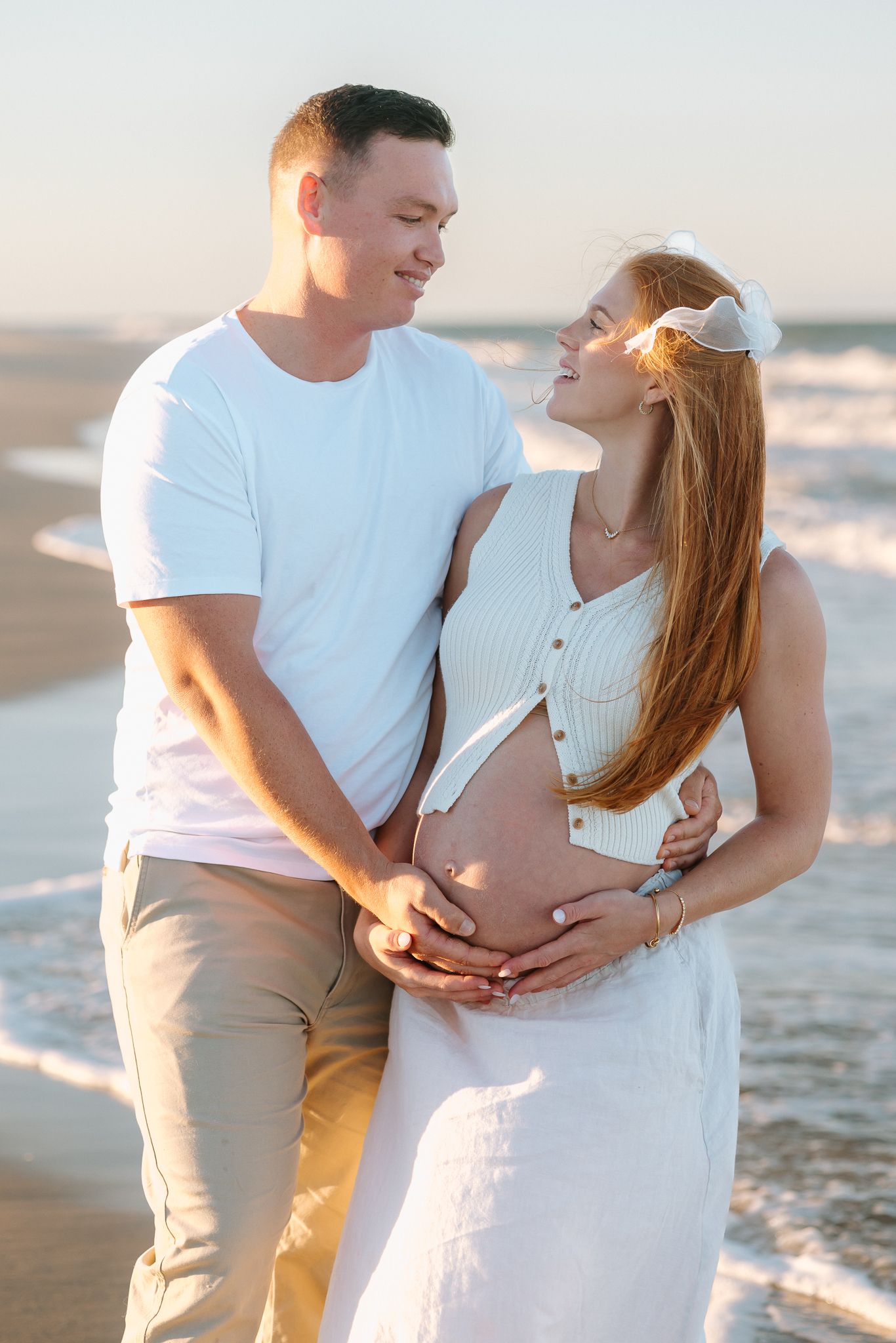 Couple embracing on beach at sunset, woman in white dress showing pregnant belly, man in white shirt holding her bump.