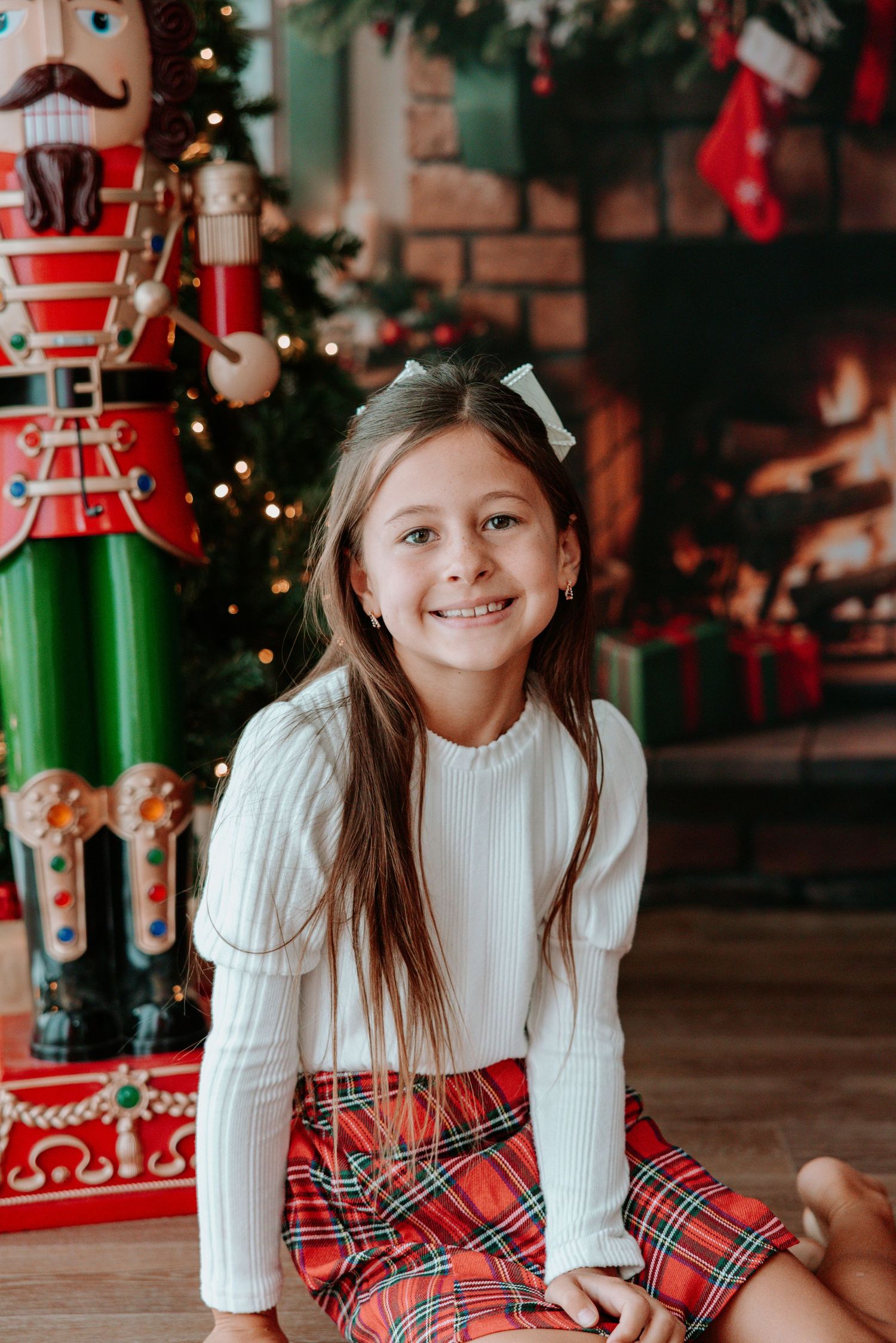 Smiling girl in white sweater and plaid skirt sits before a Christmas tree with nutcracker and fireplace.