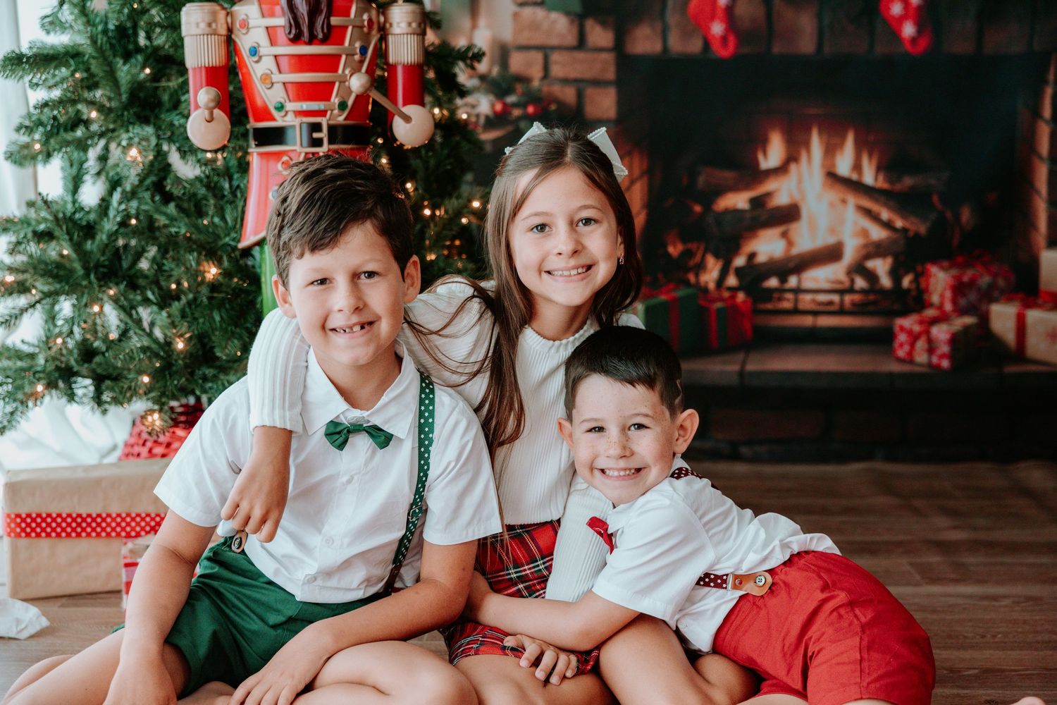 Three children in Christmas outfits smile together near a decorated tree and fireplace.