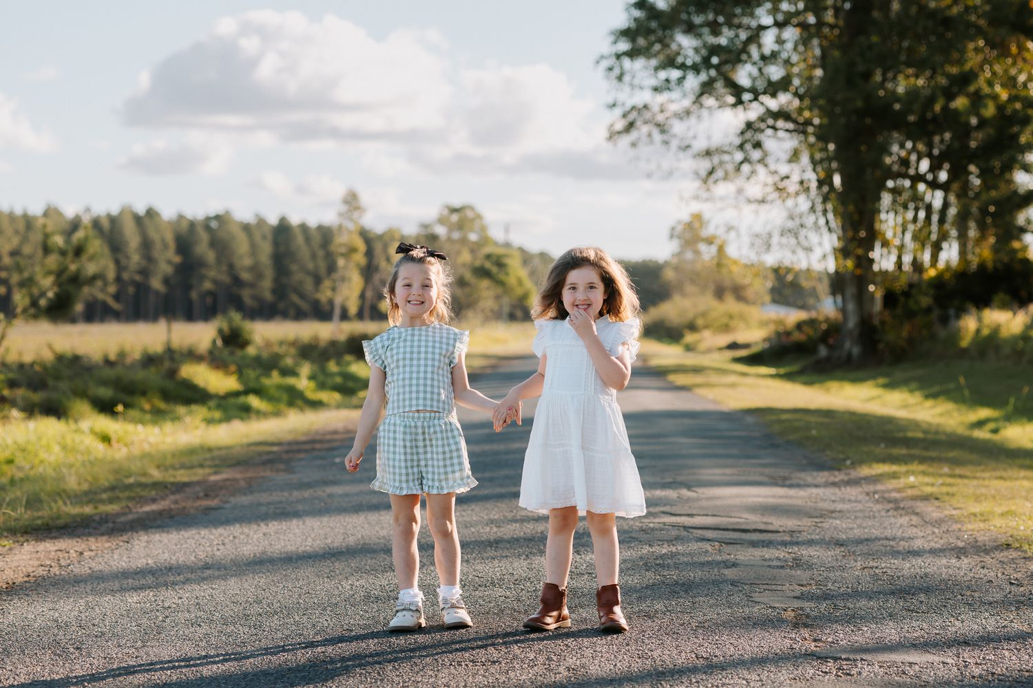 Two young girls in dresses holding hands on a rural road surrounded by trees and green fields on a sunny day.