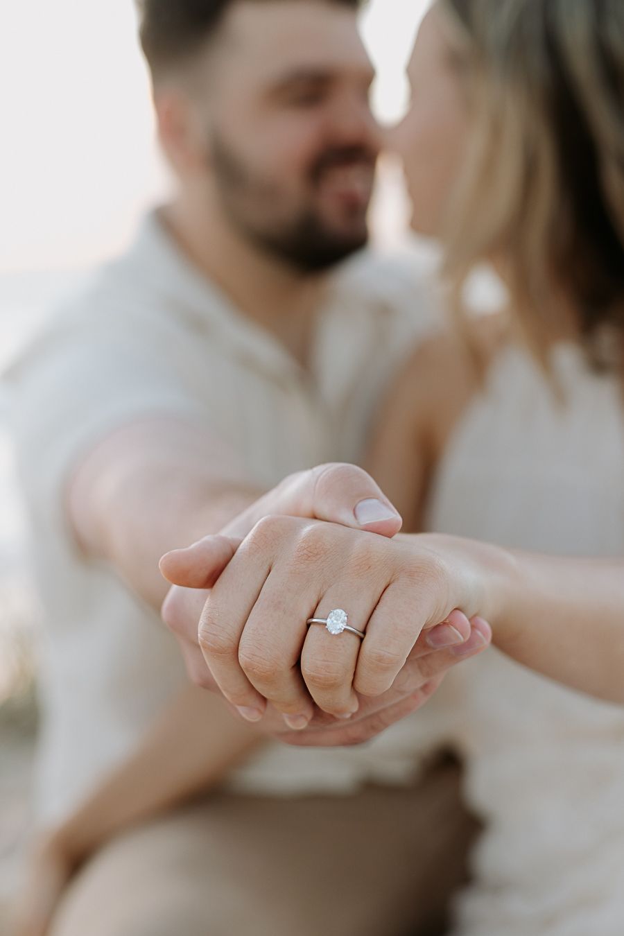Couple holding hands showing oval diamond engagement ring, man and woman in white outfits sharing romantic moment.