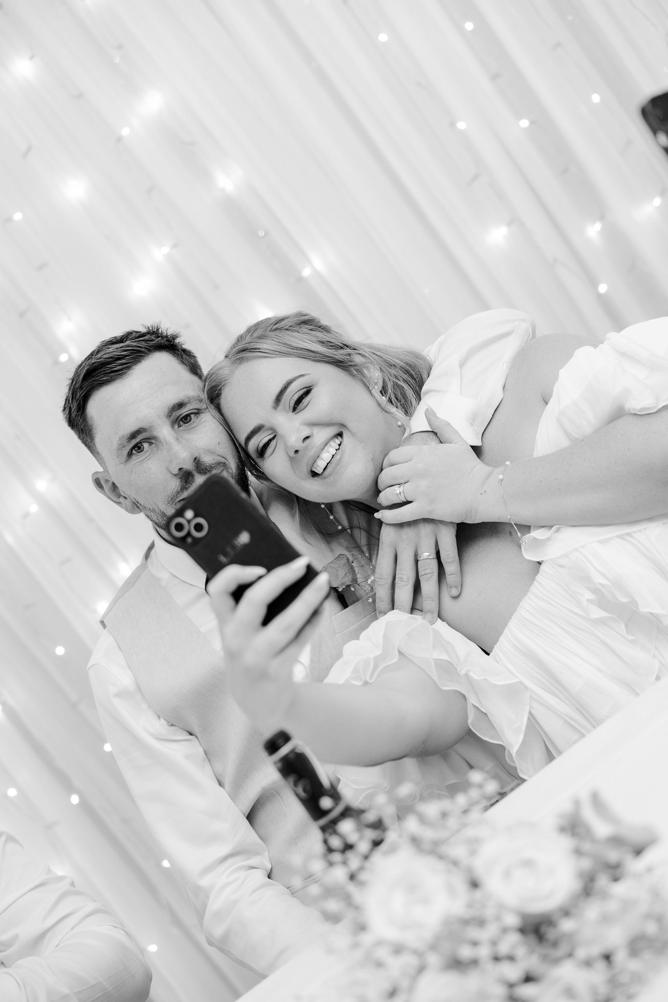Black and white photo of smiling bride and groom taking a selfie at their wedding reception.