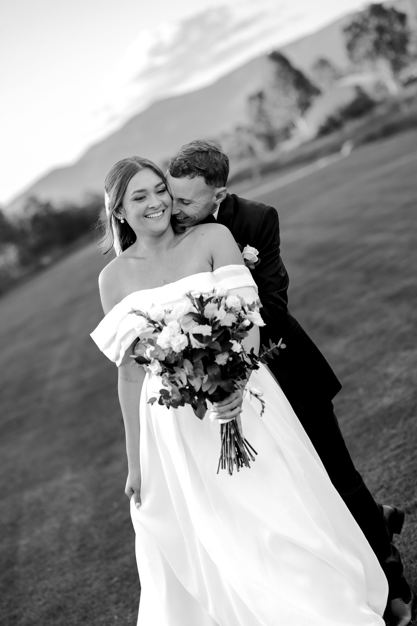 Black and white wedding photo of groom embracing bride holding bouquet outdoors with mountain backdrop.
