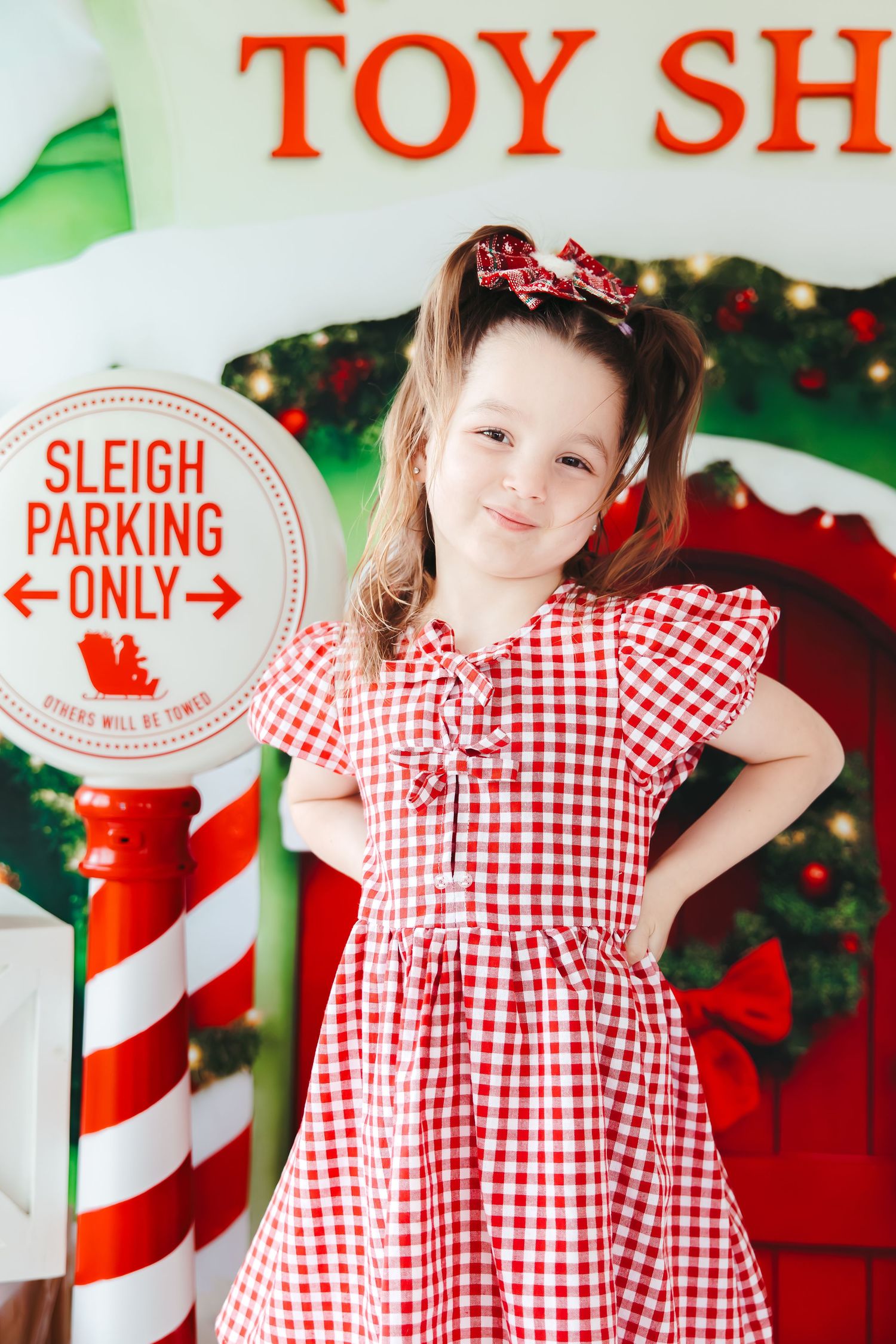 Young girl in red gingham dress poses by Christmas toy shop sign with sleigh parking only backdrop.