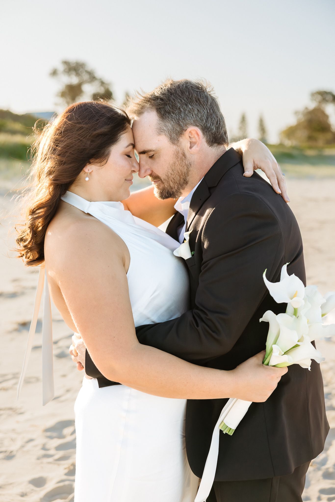 Bride and groom share a romantic forehead touch on a sandy beach, she holds white calla lilies in a halter gown.