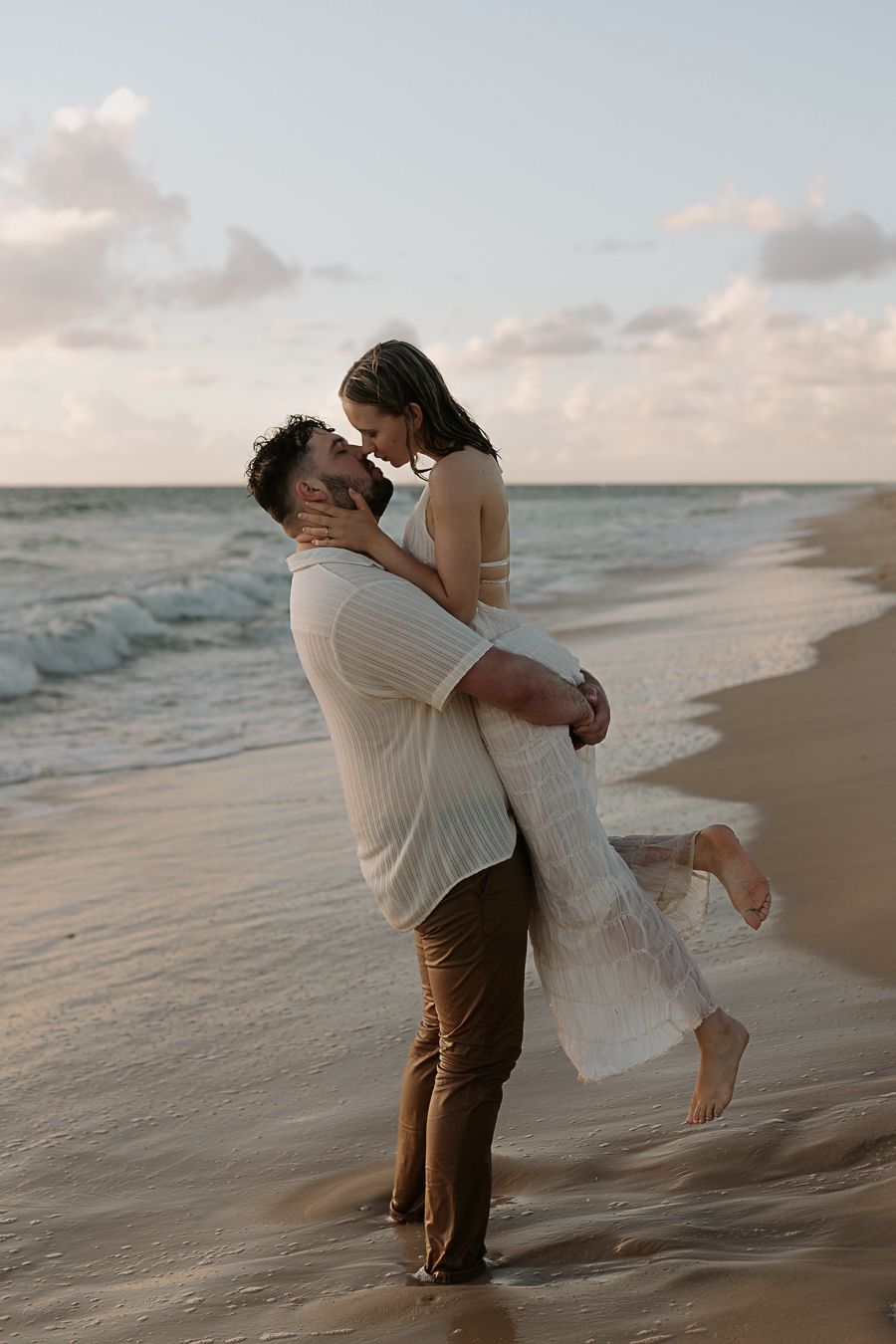 Romantic couple embracing on a beach at sunset, man lifting woman in a white dress by the ocean.