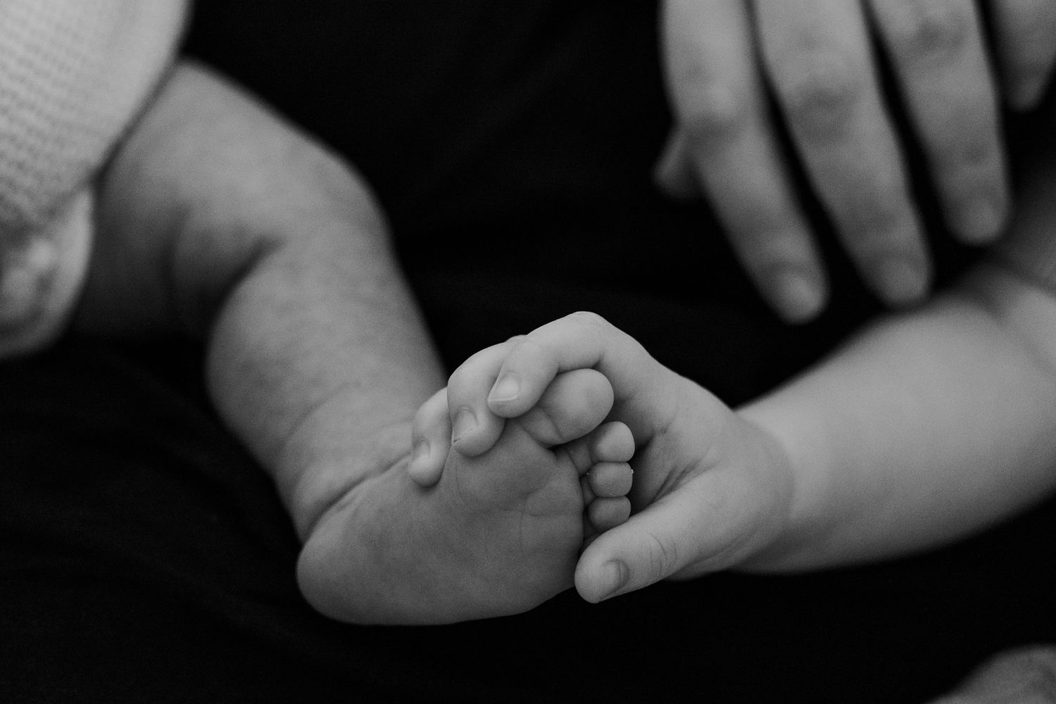 Black and white close-up of a newborn baby's tiny feet cradled gently in adult hands.