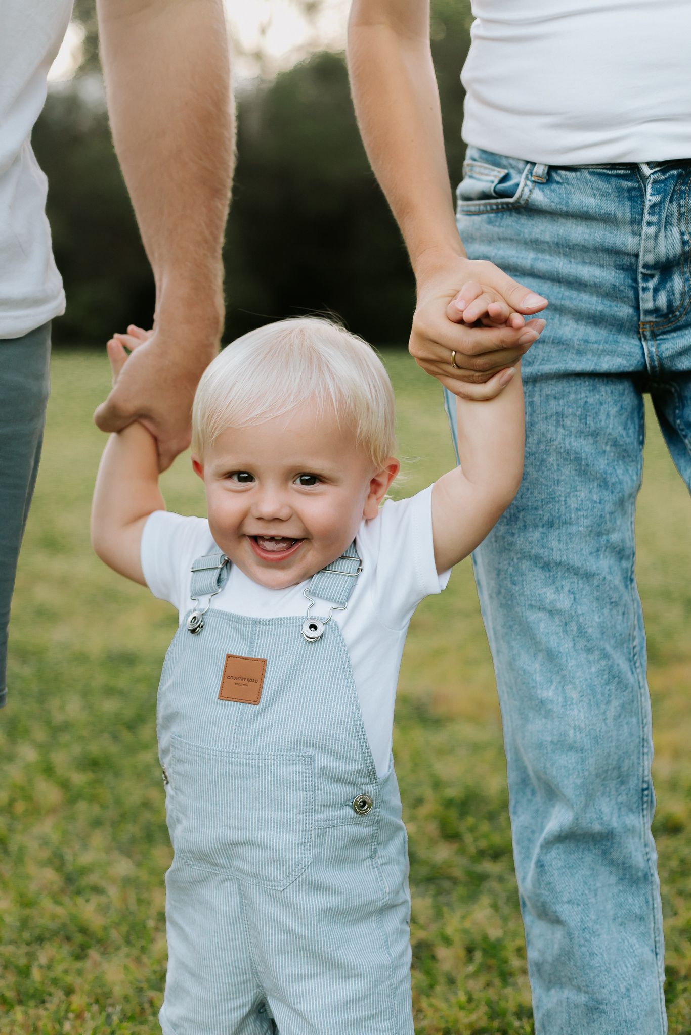 Smiling toddler in overalls holds parents hands while standing on green grass outdoors.