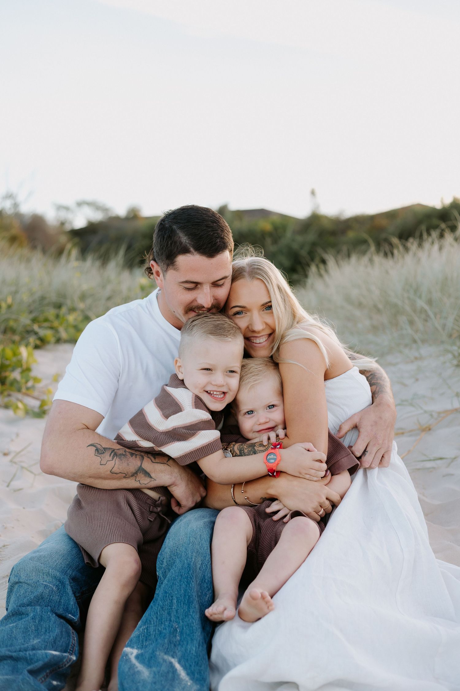 Happy family of four sharing a warm embrace on a sandy beach, with lush green dunes in the background.