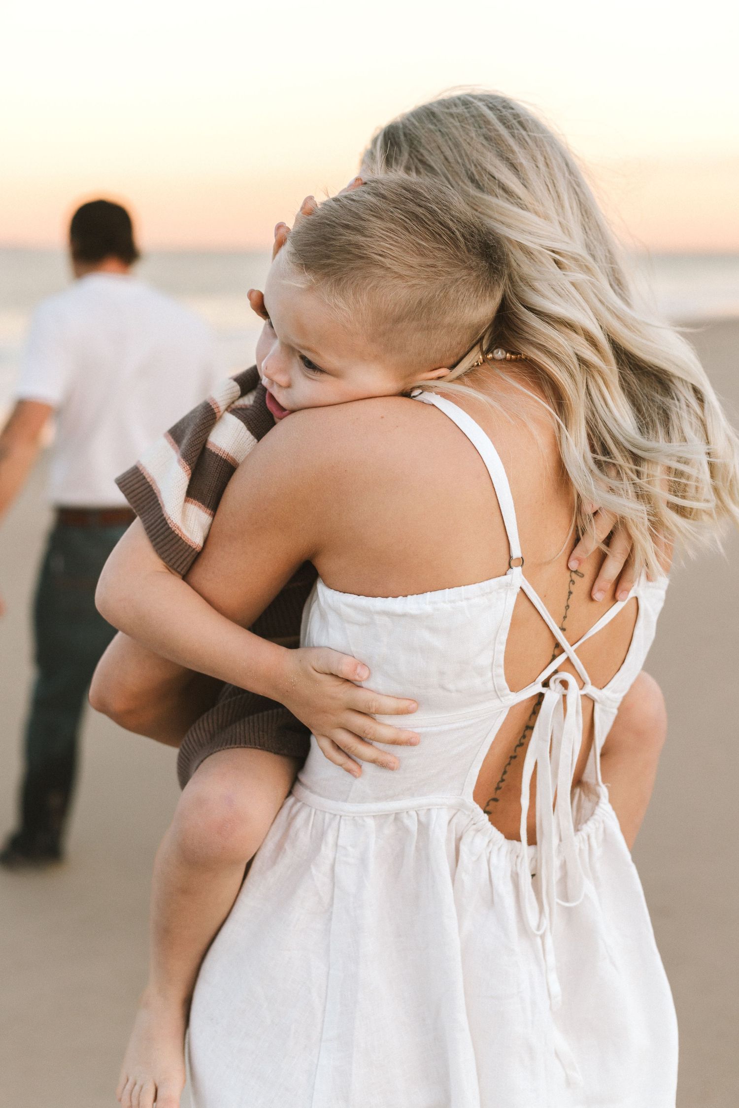 Woman in white sundress holding child on beach at sunset with man walking in background.