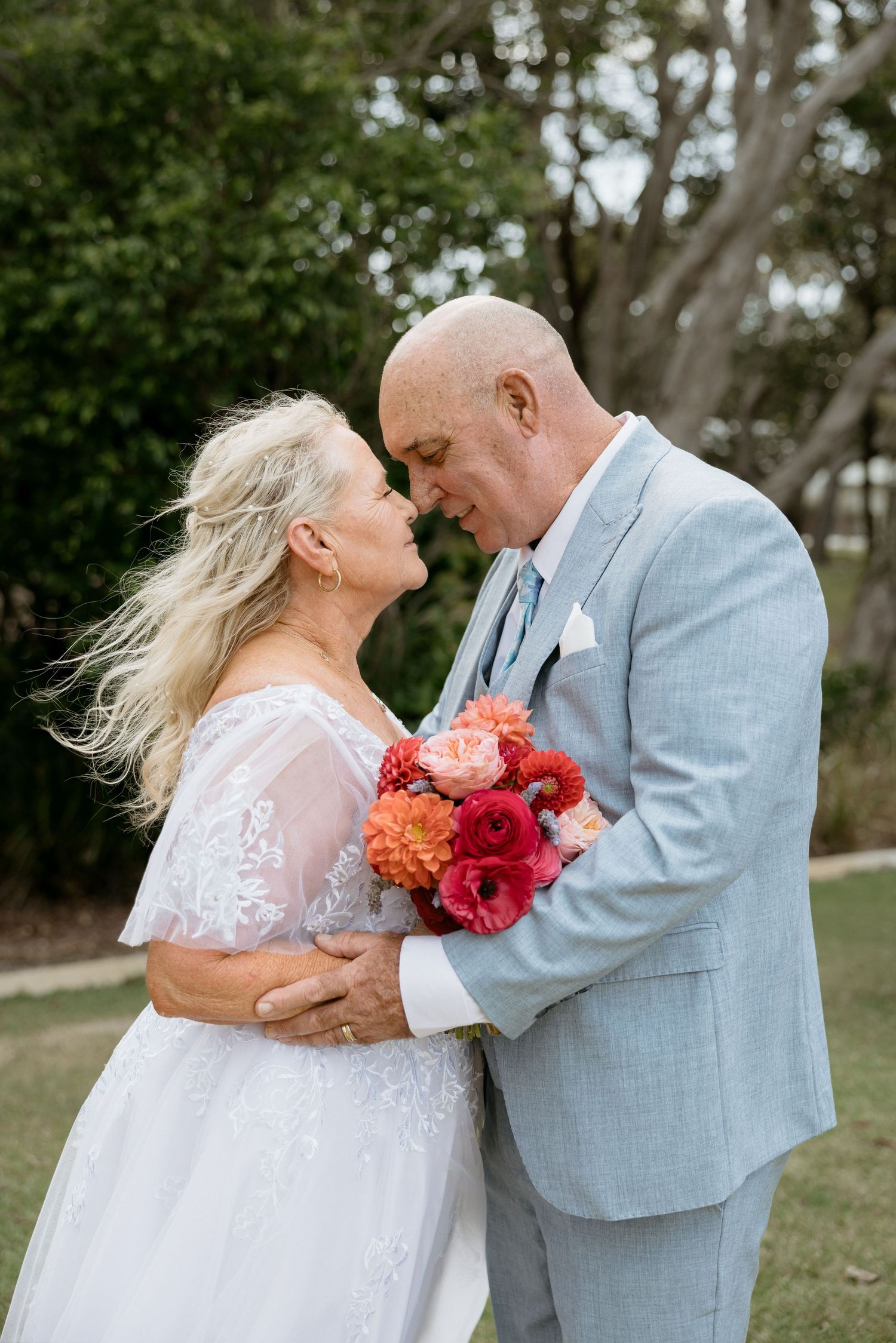 A couple in wedding attire share intimate moments in a garden setting with a vibrant coral and red bouquet between them.