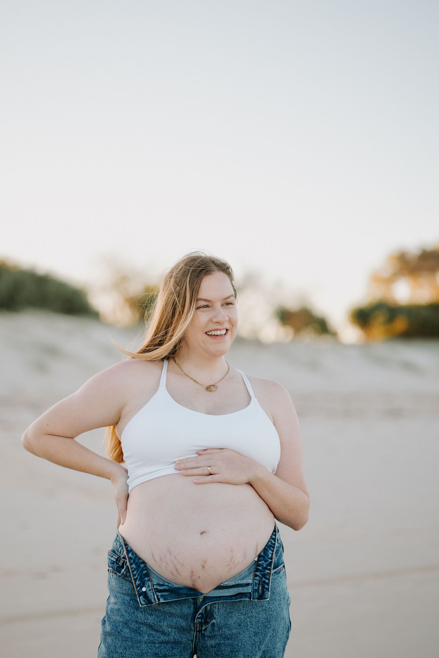 A person in jeans and a white tank top laughs joyfully while sitting on a sandy beach at sunset.