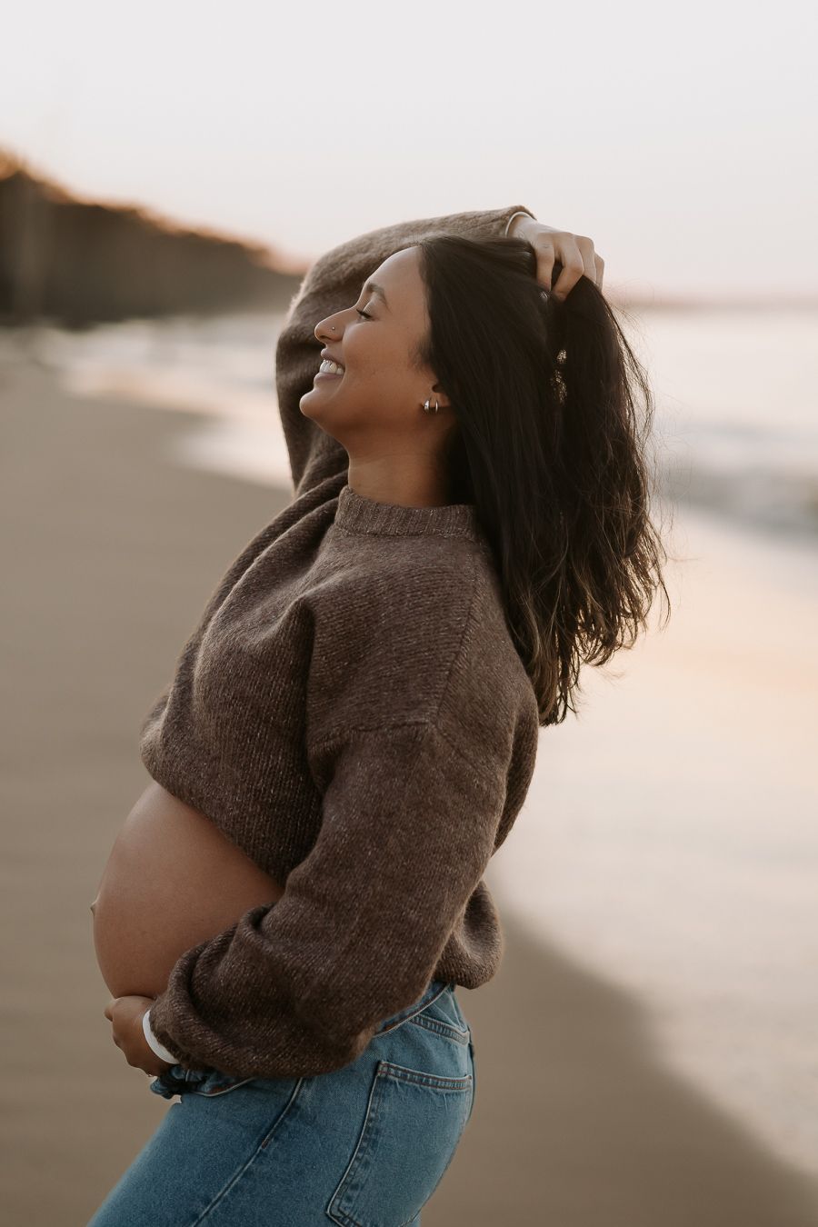 A person in a brown sweater and jeans poses expressively against a sunset beach backdrop.