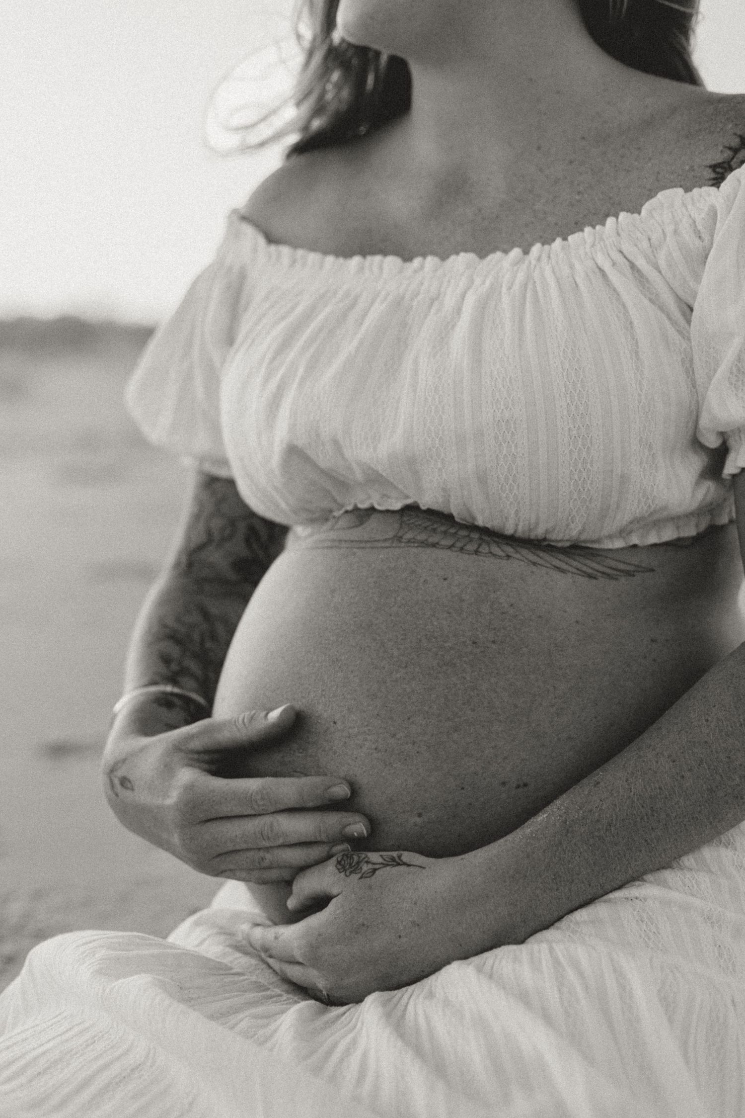 A black and white photo shows a pregnant belly being cradled by hands, with a flowing white off-shoulder dress.