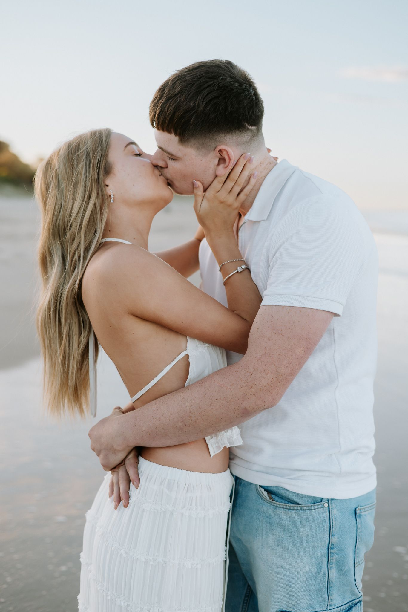 Couple sharing a romantic kiss on the beach at sunset, woman in white backless dress embracing man.