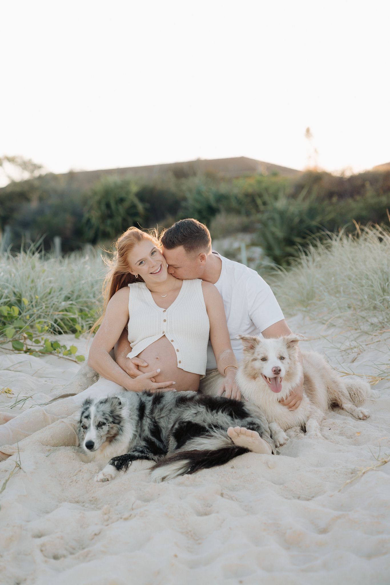 Pregnant couple sitting on sandy beach with two dogs at sunset, man kissing womans cheek, wearing white outfits.