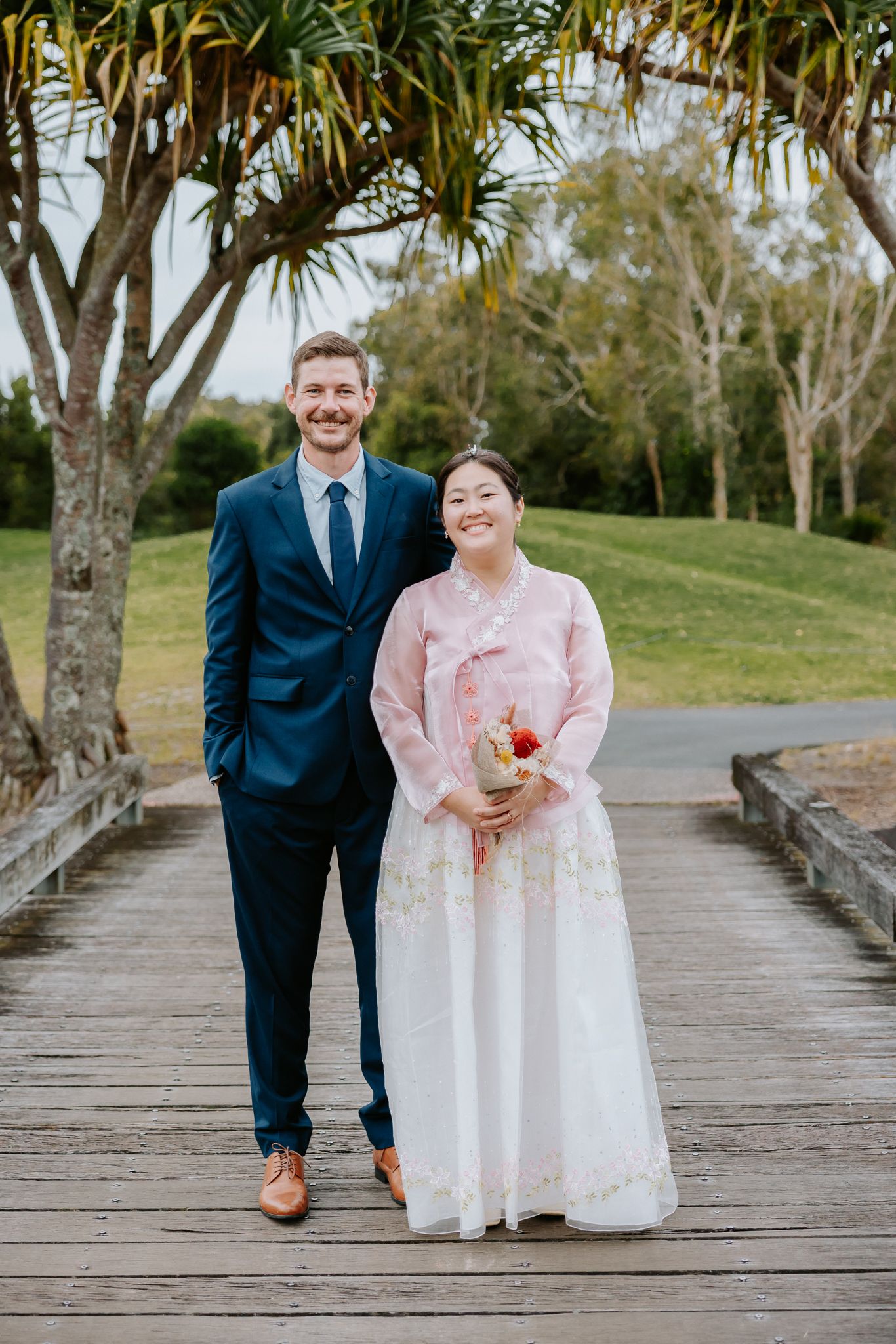 Couple posing on wooden bridge, man in navy suit, woman in pink Korean hanbok holding bouquet, outdoor park setting.