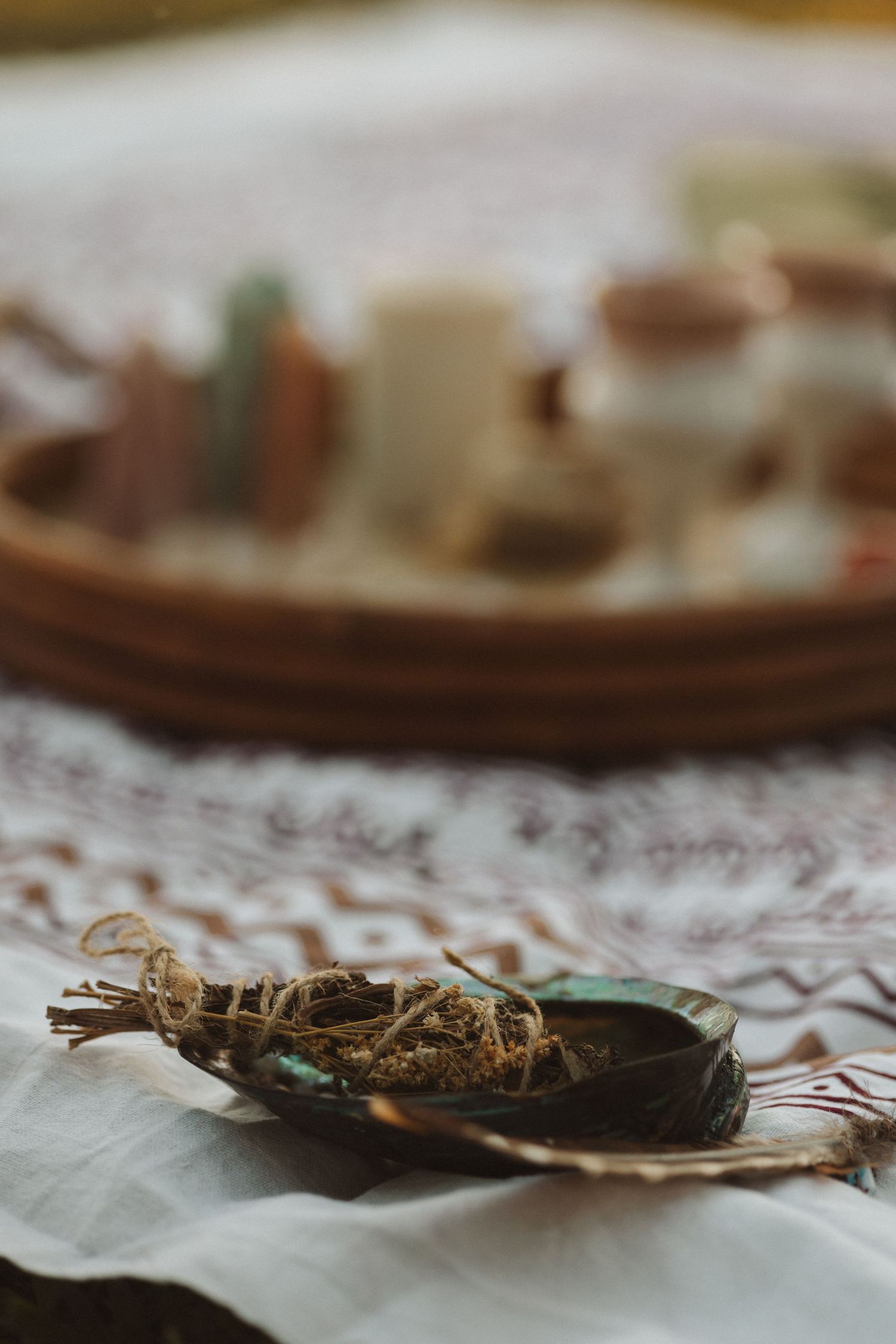A sage smudge stick resting on an abalone shell on embroidered cloth, with a blurred basket of crystals behind.