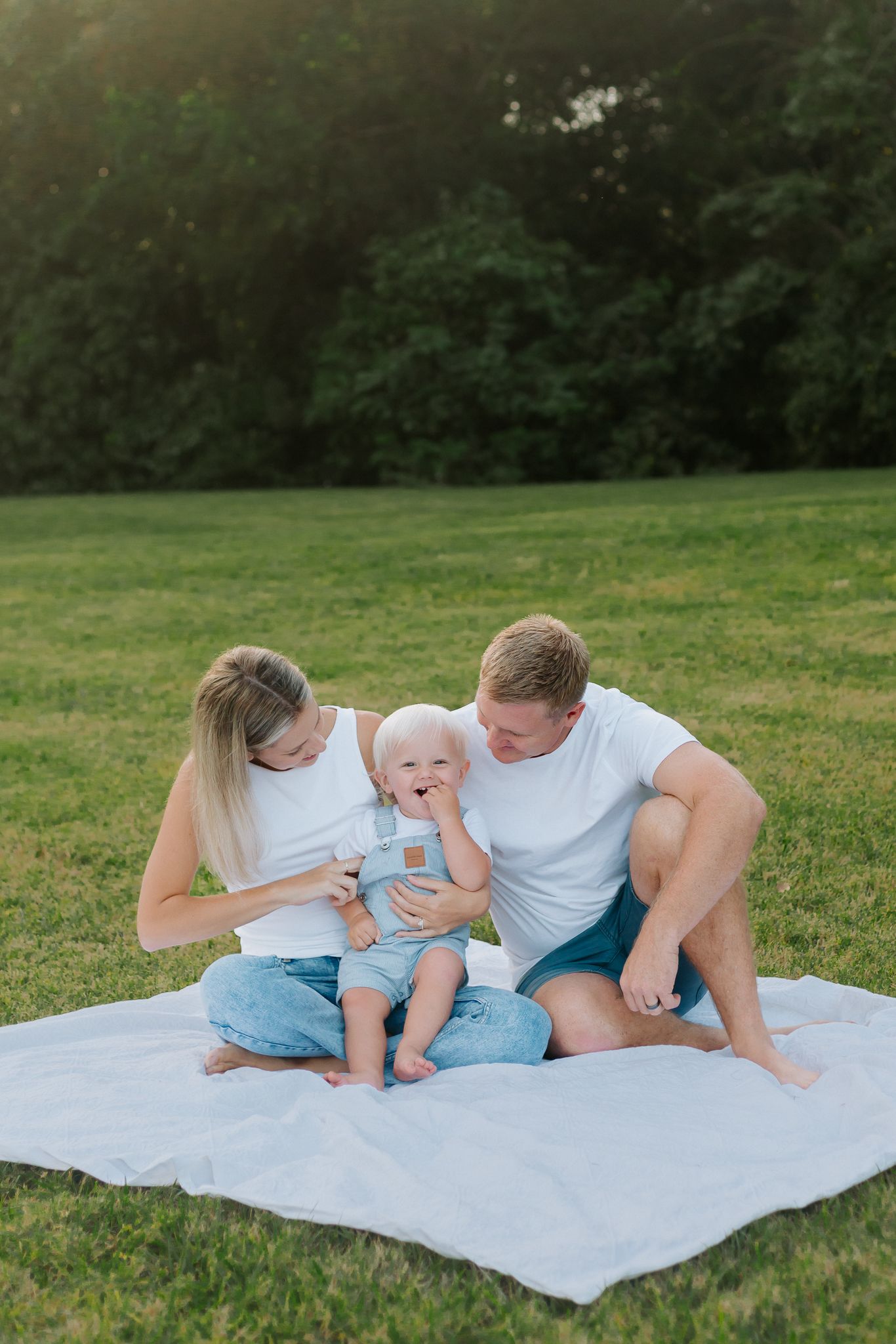 Young family with baby sitting on white blanket in green park during golden hour sunset.