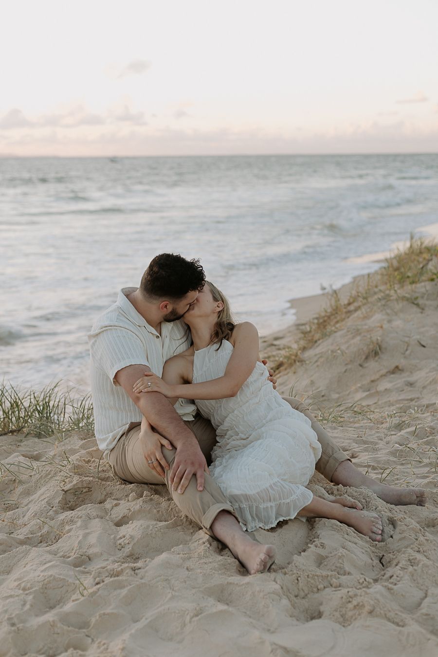 Romantic couple sitting together on beach sand dunes at dusk, sharing a kiss near the ocean waves.