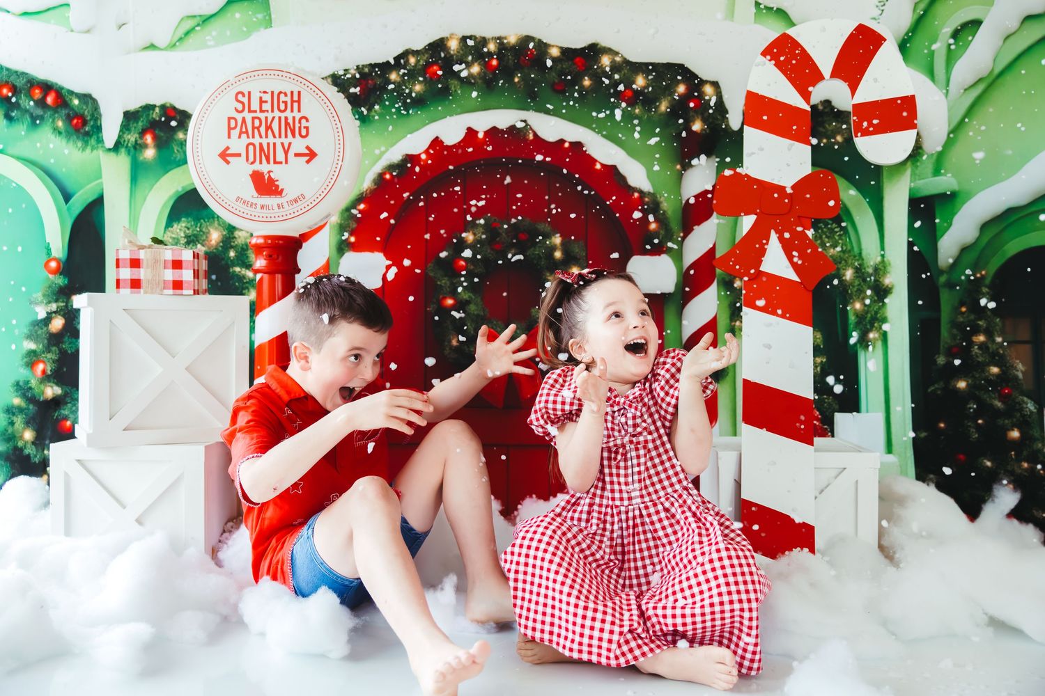Two children laugh and play in a festive Christmas photo set with candy canes, a sleigh parking sign, and holiday decorations.