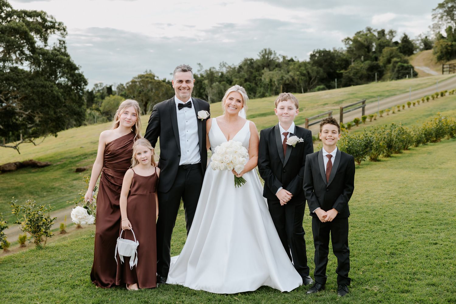 Wedding couple poses with family including bridesmaid and children on lush green lawn during outdoor ceremony.