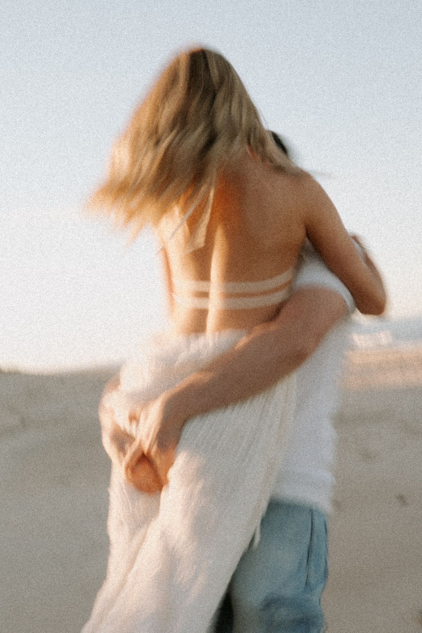 Blurred motion shot of a blonde woman in a white backless top twirling on a sandy beach at golden hour.
