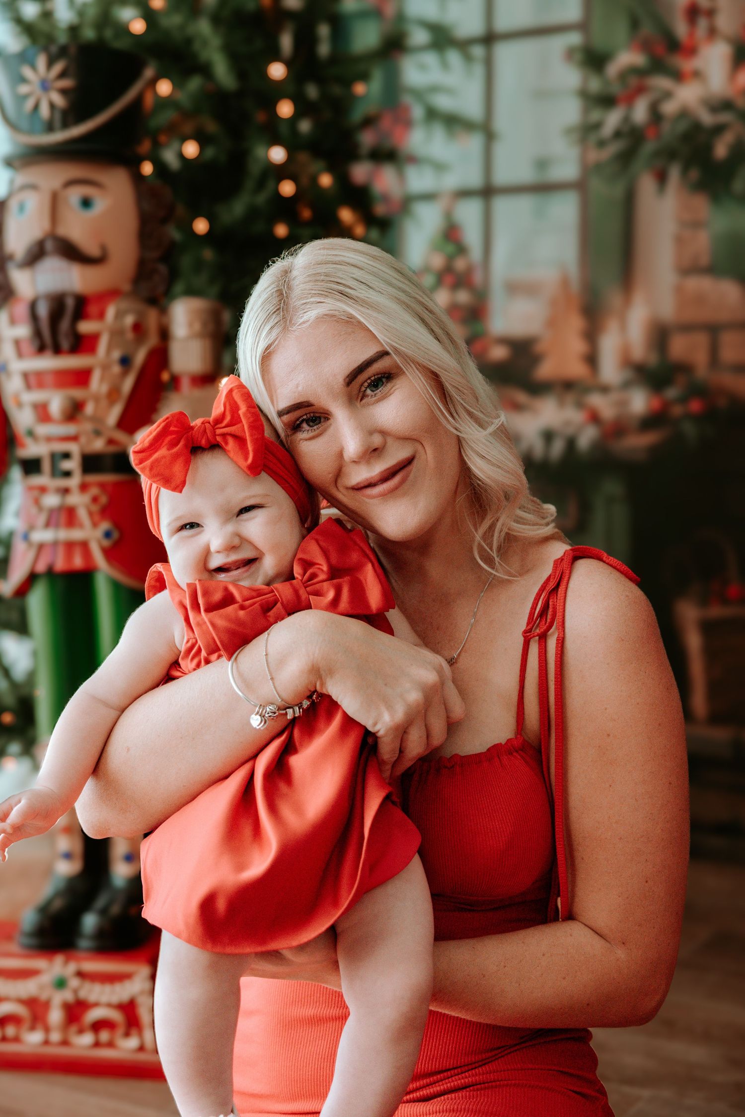 Woman in red dress holding smiling baby in red outfit in front of Christmas tree with nutcracker decor.