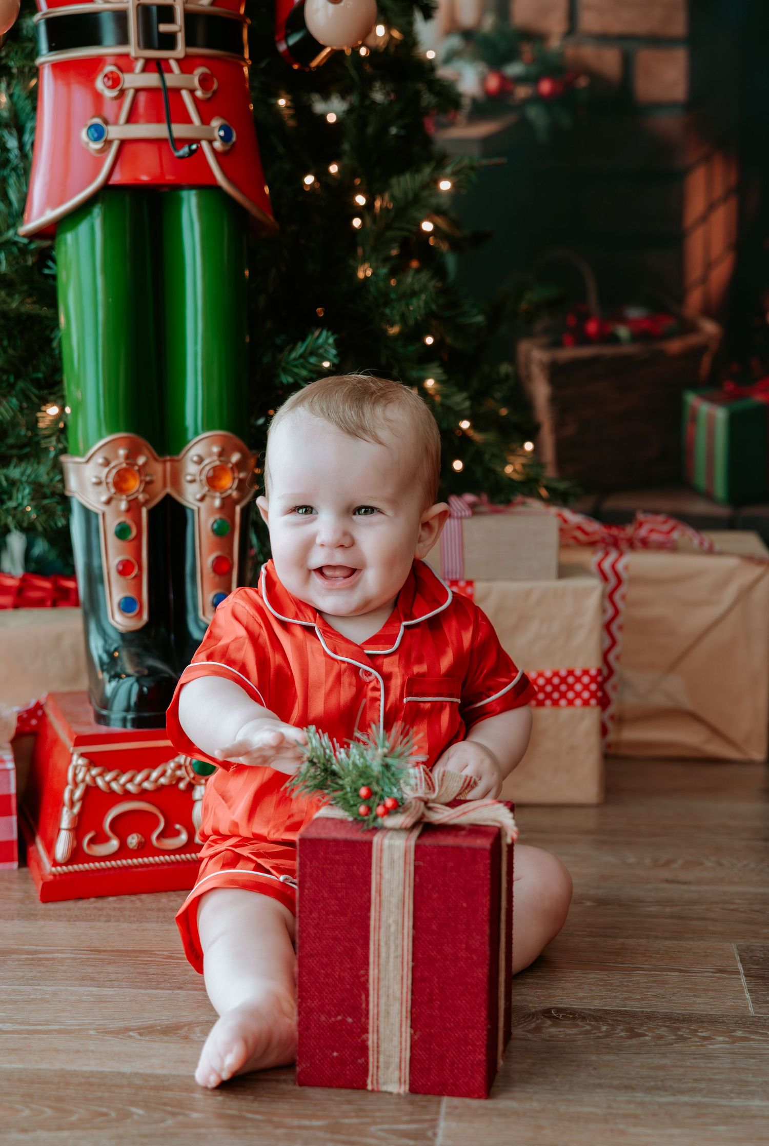 Baby in red pajamas smiling while opening a Christmas gift, surrounded by presents and a decorated tree.