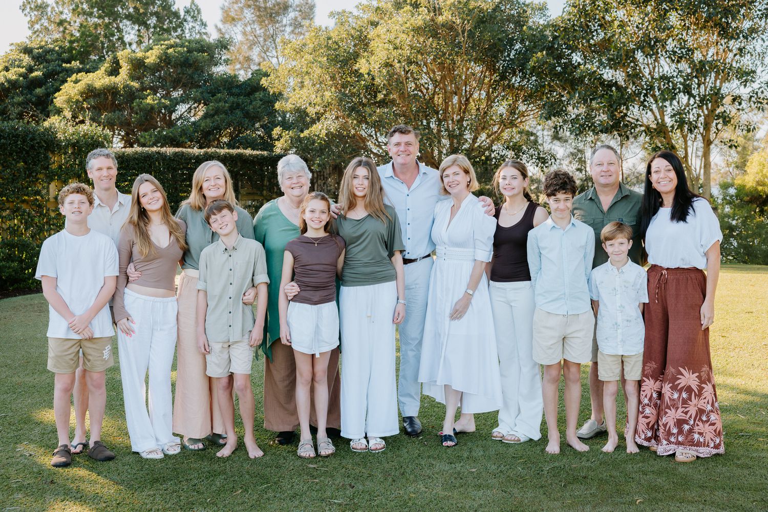 Large multigenerational family posing together outdoors on green lawn with lush trees in background during daytime.