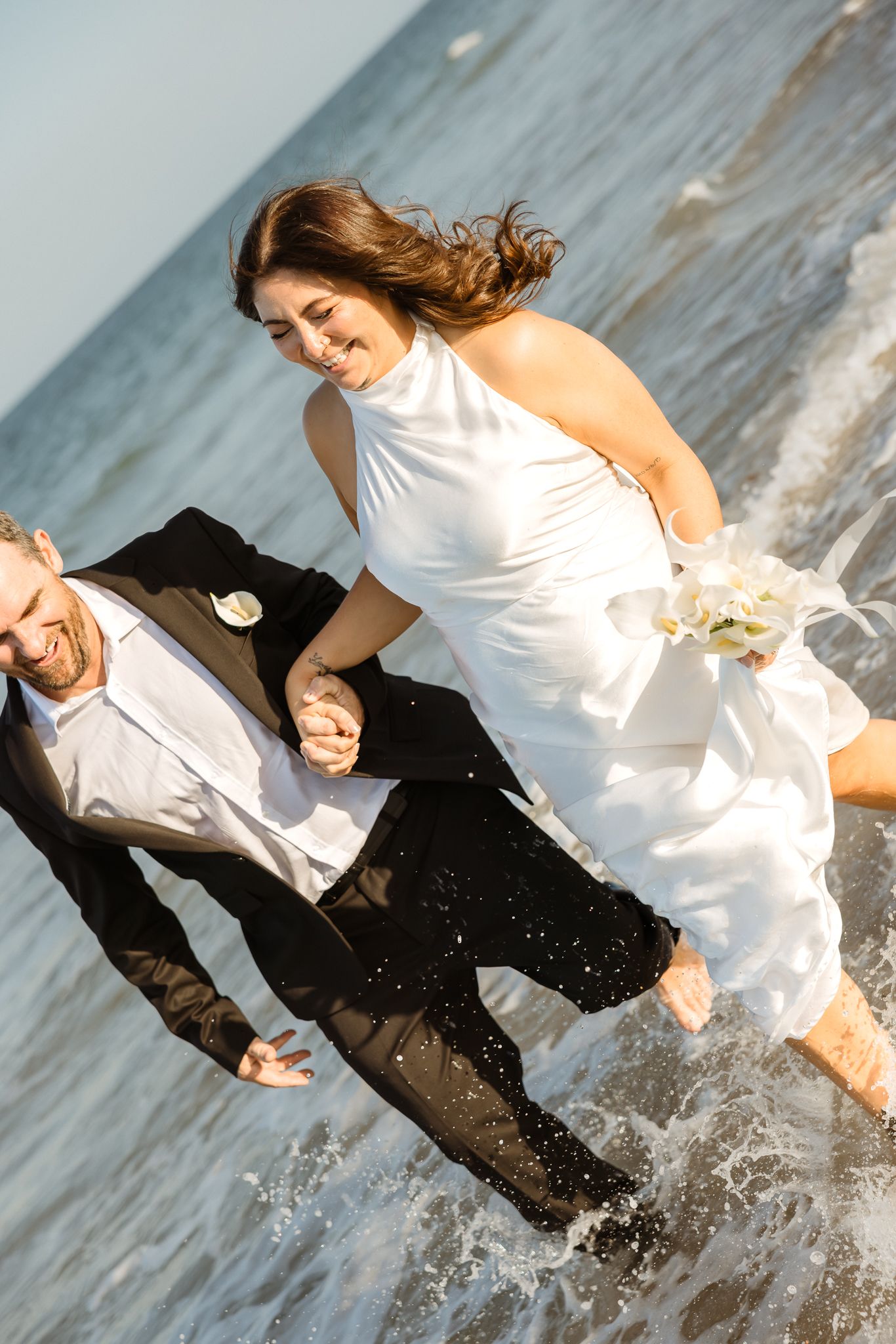 Joyful newlyweds splashing through ocean waves on beach, bride in white halter gown holding bouquet, groom in black suit.