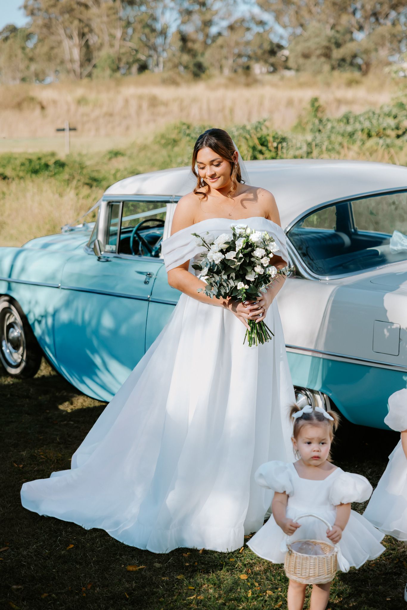 Bride in off-shoulder white gown holding bouquet beside vintage teal car, with young flower girl in white dress.