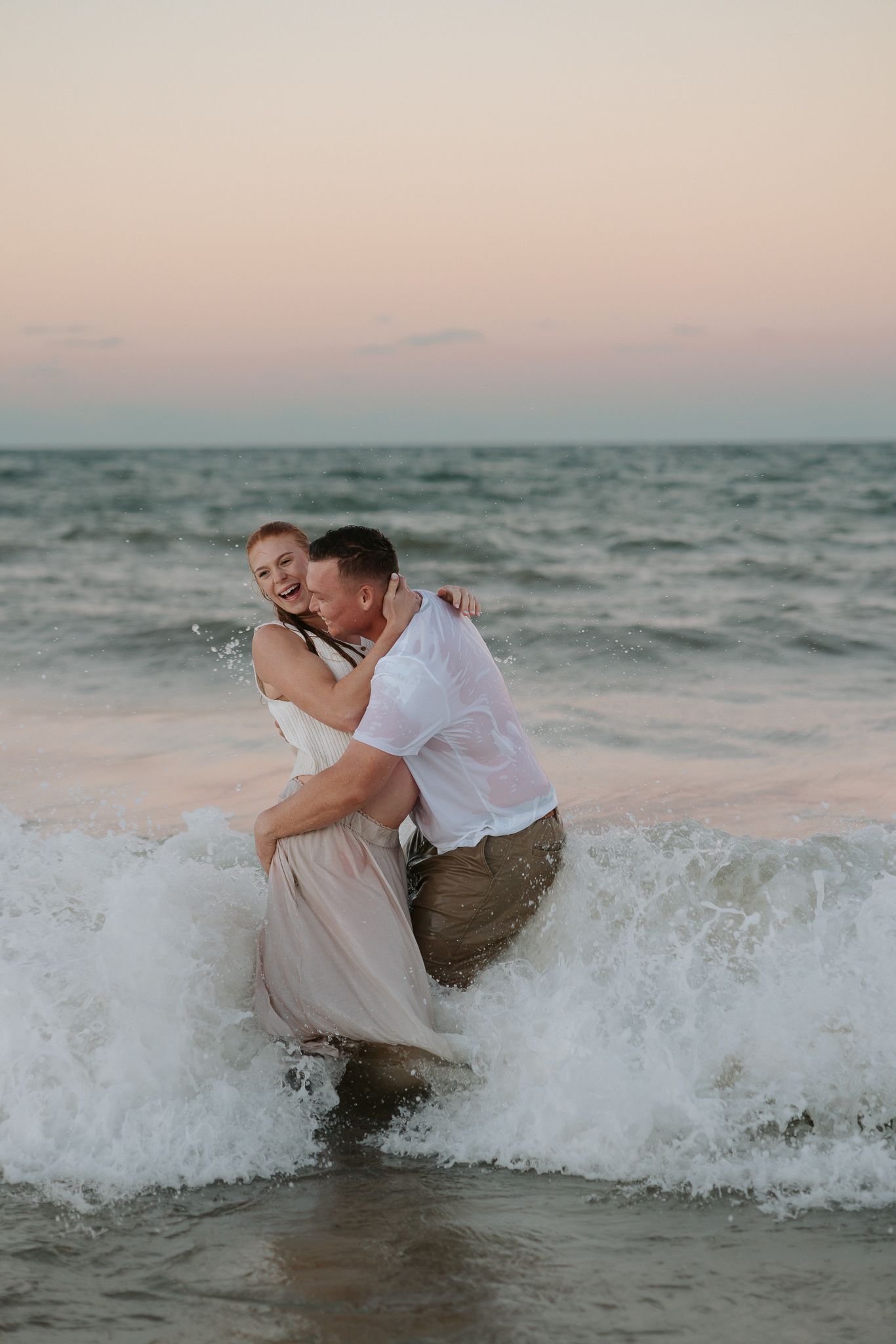 Couple embracing in ocean waves at sunset beach, romantic engagement photo with pastel sky backdrop.