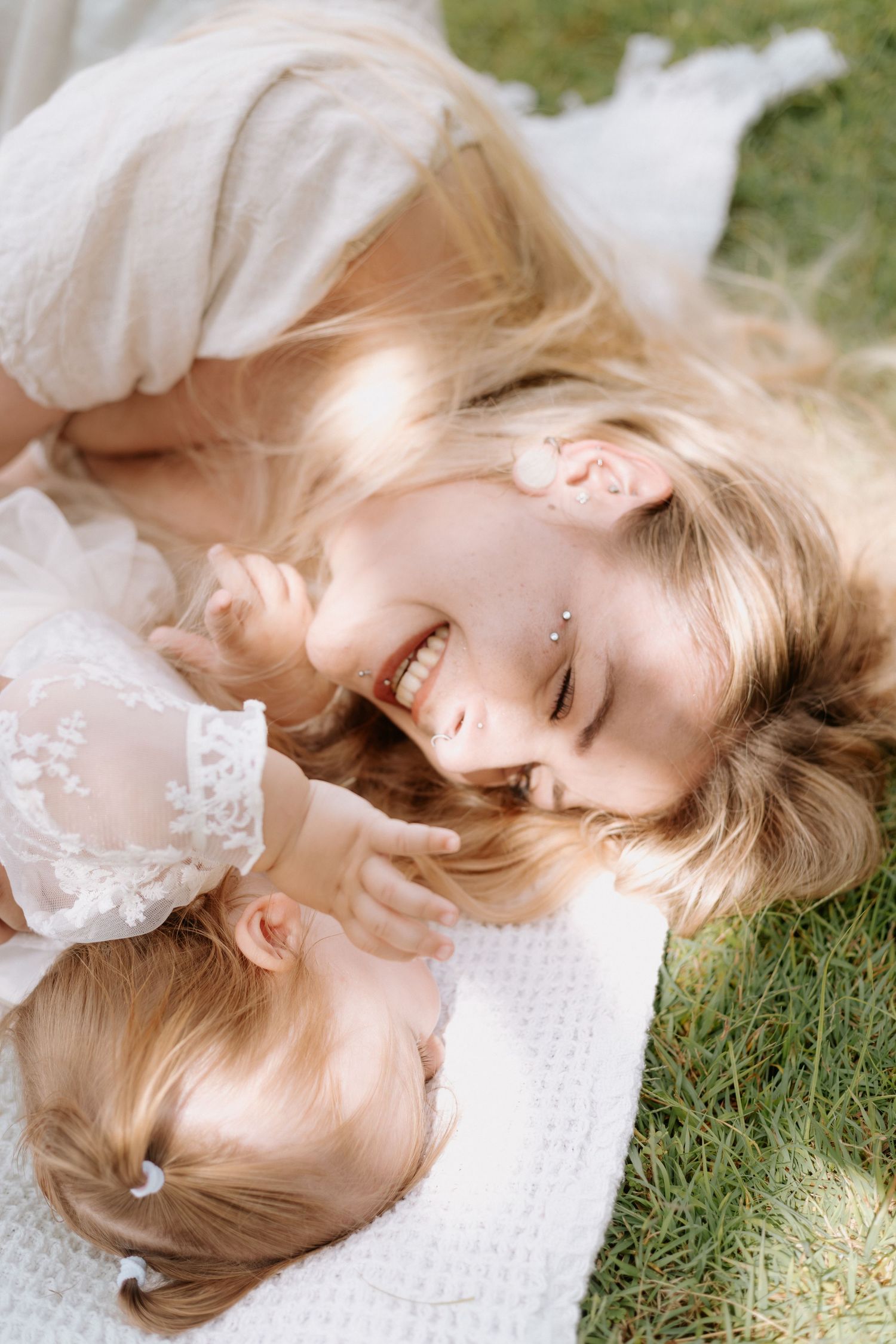 Two people in white lace dresses lay on grass together, sharing a joyful intimate moment while smiling.