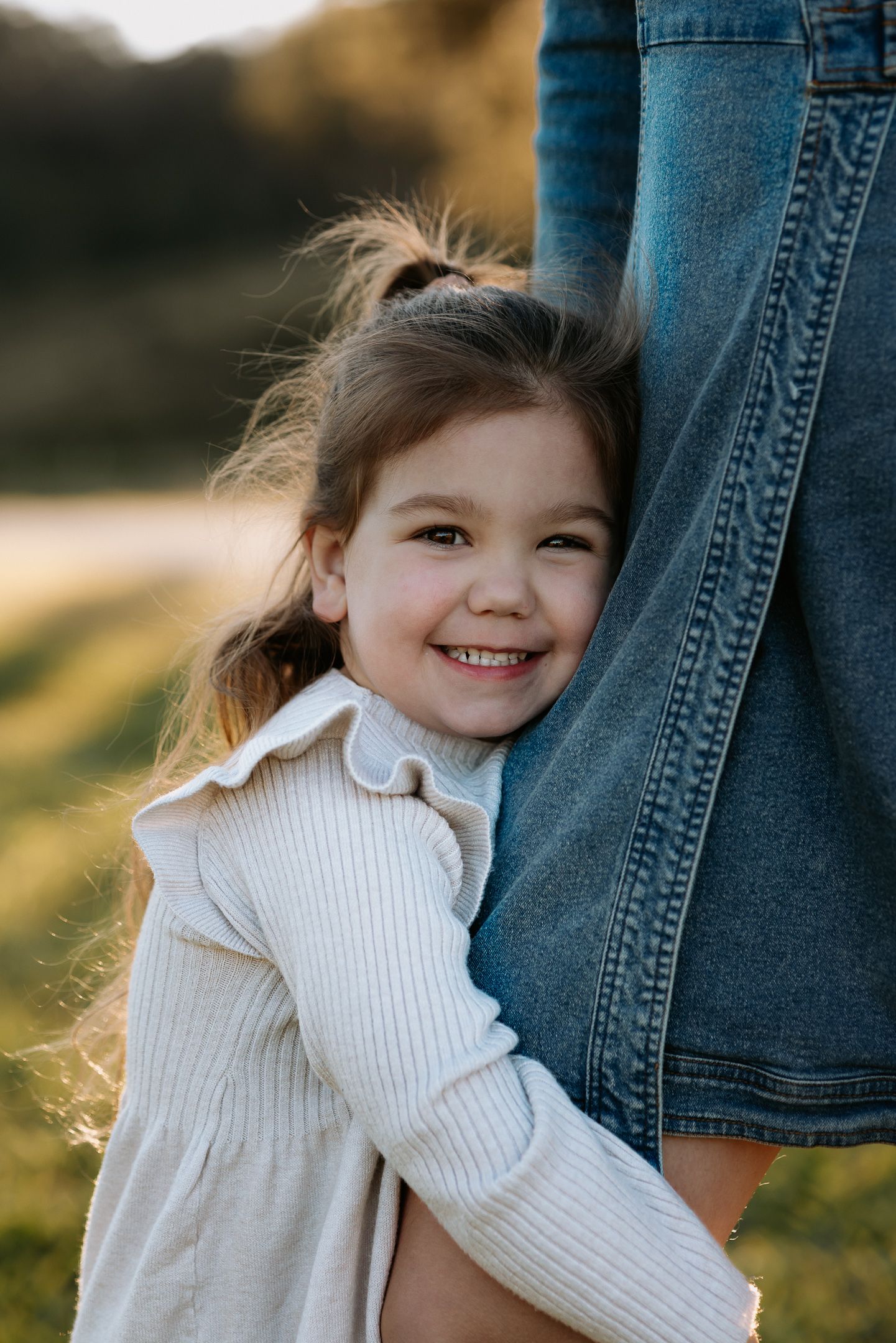 A young child in a white sweater playfully hides behind someone wearing jeans in a sunlit outdoor setting.