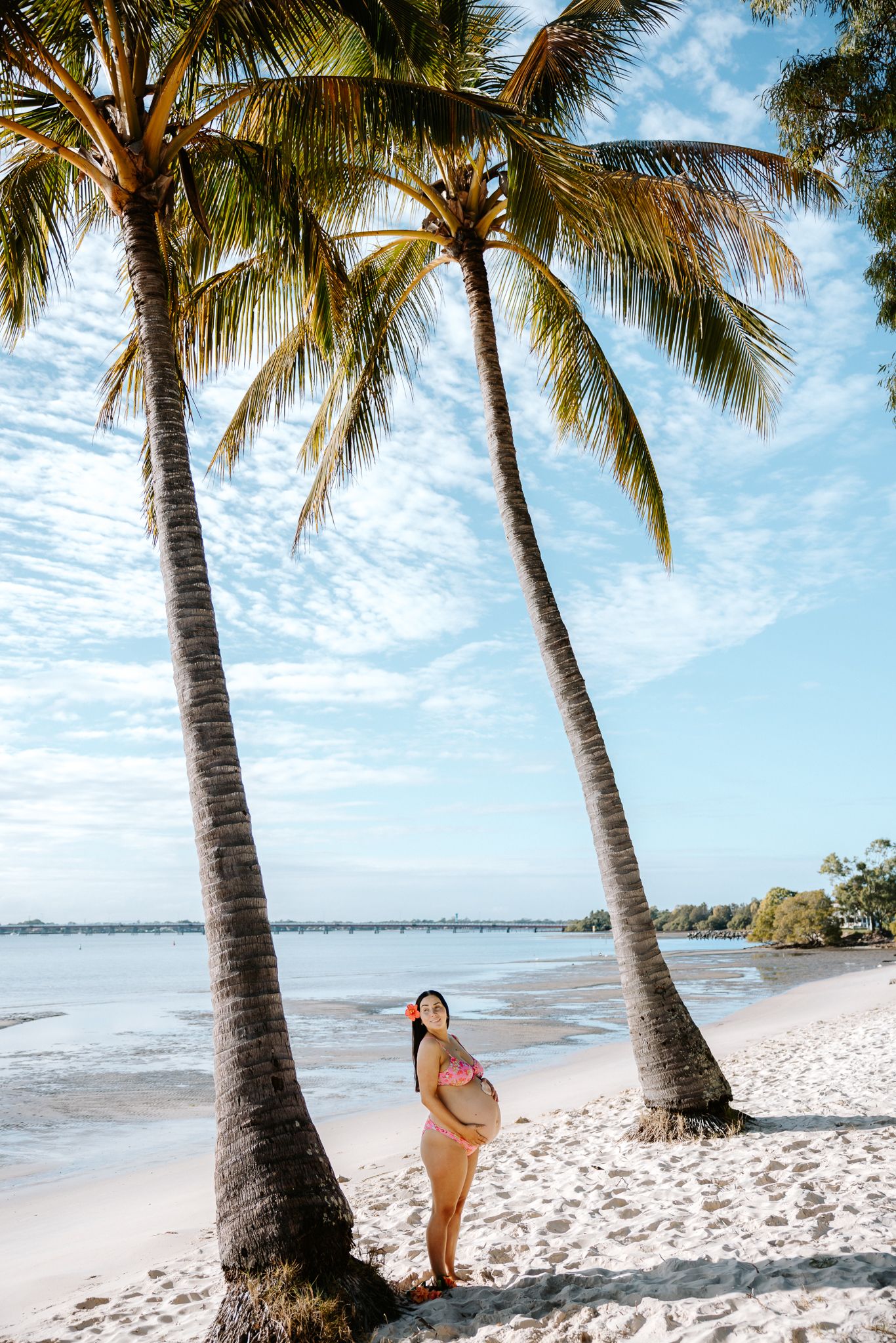 Pregnant woman in bikini standing between palm trees on a tropical white sand beach under a blue sky.