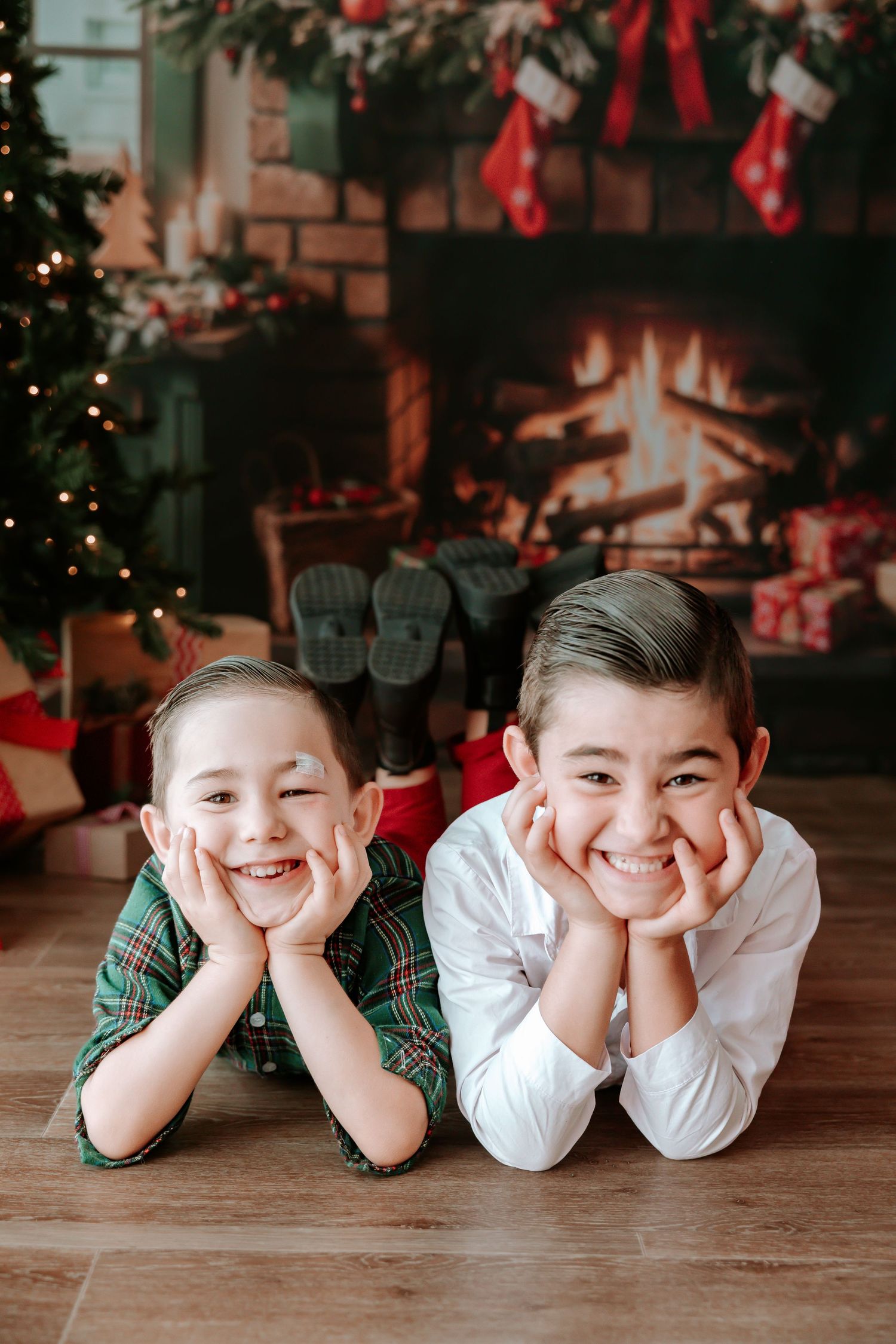 Two smiling boys lying on floor in front of decorated Christmas fireplace with stockings and tree.