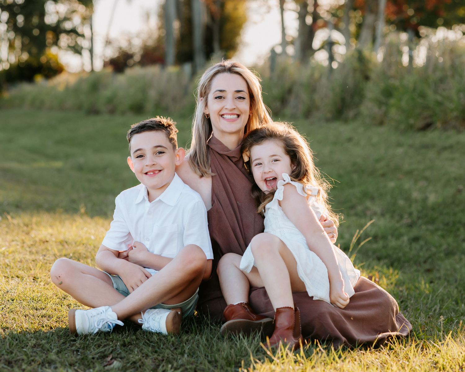 A smiling woman sits on grass with two happy children in a sunny outdoor family portrait.