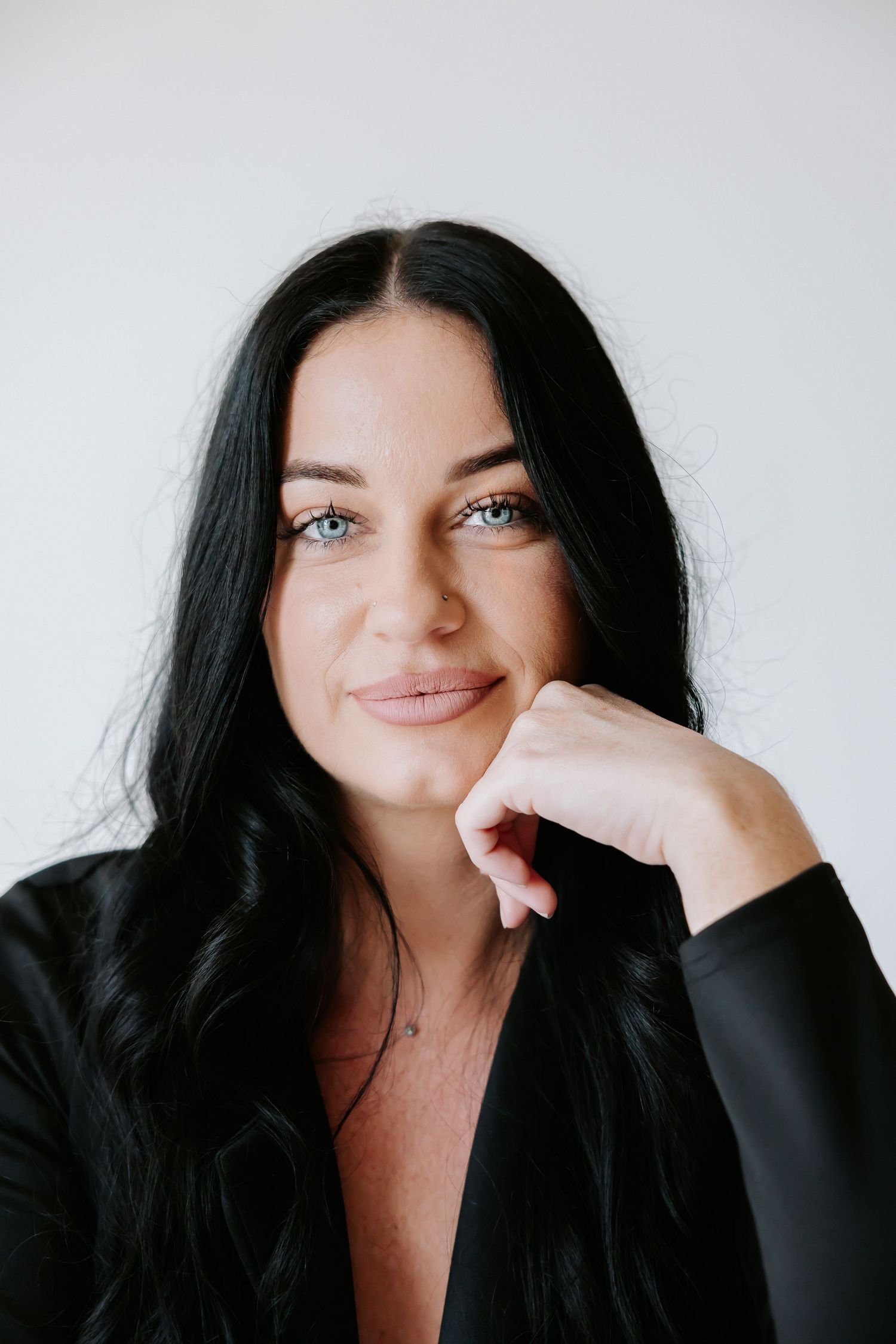 Dark-haired woman in black blazer smiling warmly with hand on chin against white background.