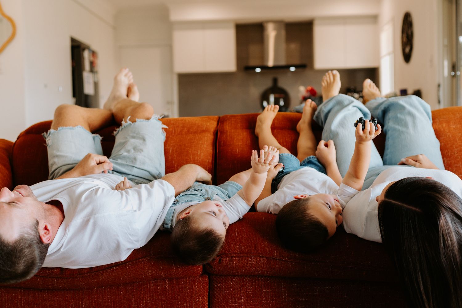Family of four lying on red sofa with feet up, laughing and relaxing together in a cozy living room.
